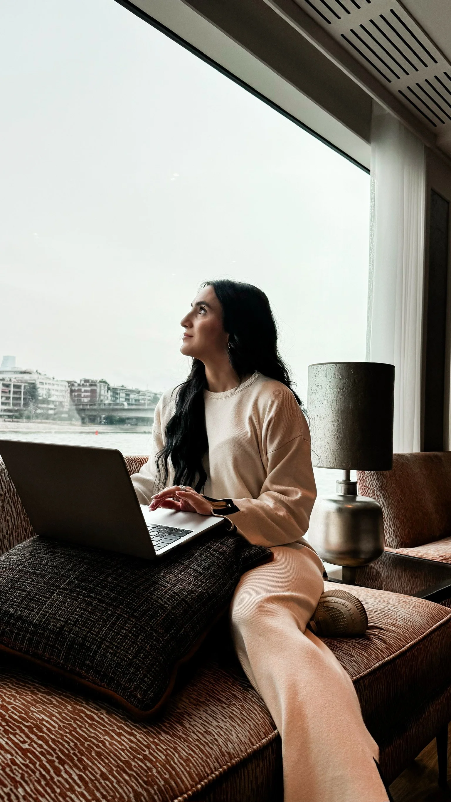 A woman in a cream-colored outfit sitting on a sofa by a large window, using a laptop with a river and cityscape outside, next to a modern table lamp.
