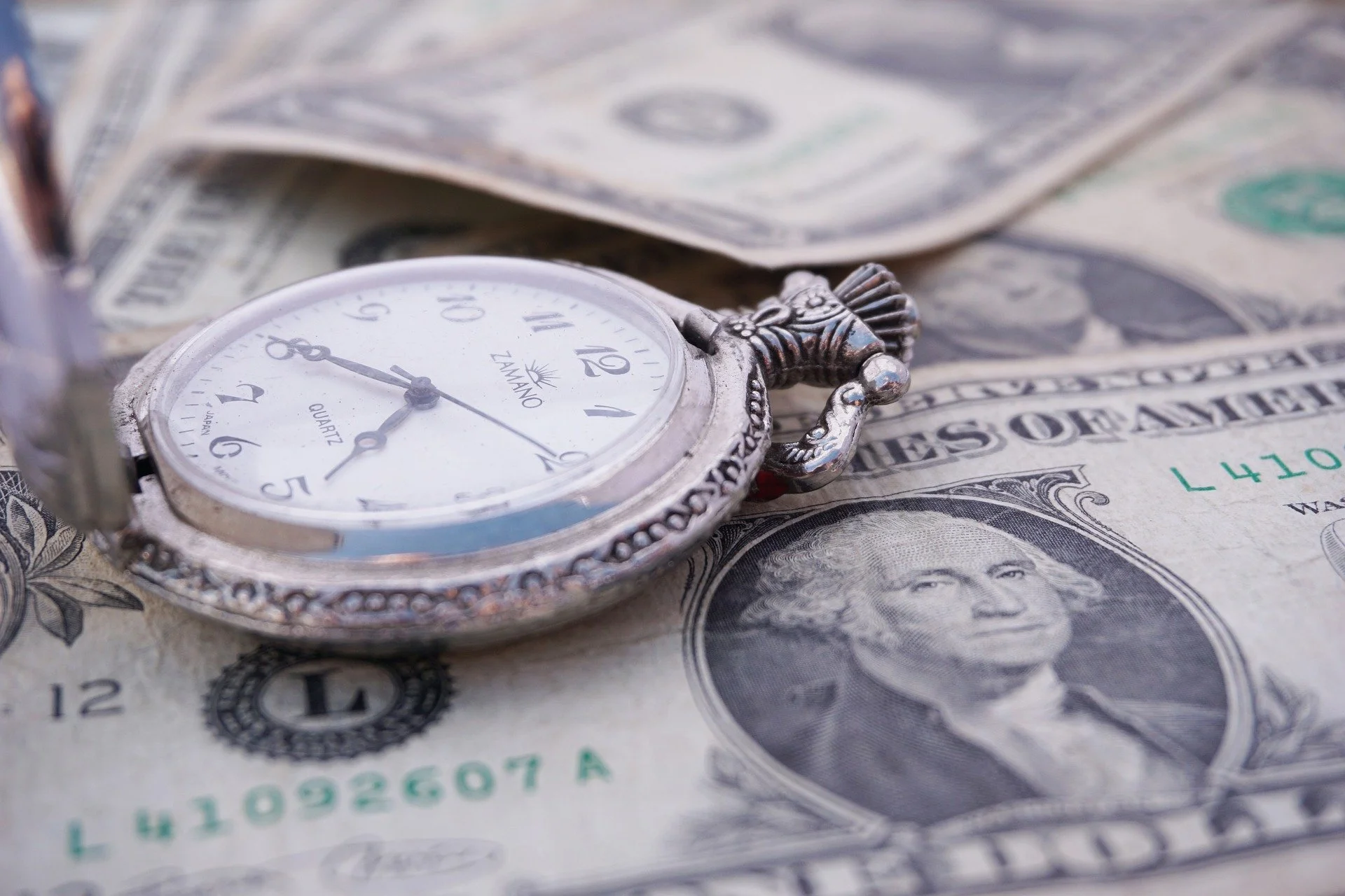 A vintage silver pocket watch with a white face lying on top of U.S. dollar bills, including a one-dollar bill with George Washington's portrait. The pocket watch shows the time as approximately 8:44.