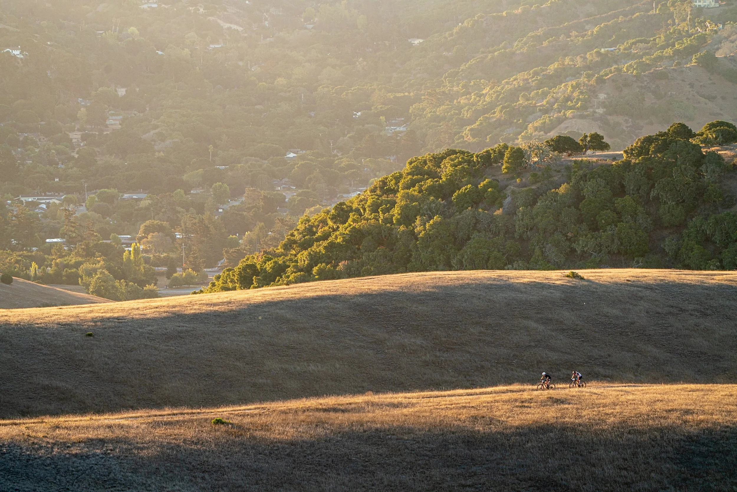 Two people riding bikes on a grassy hillside in the afternoon with a backdrop of lush green trees and hills.