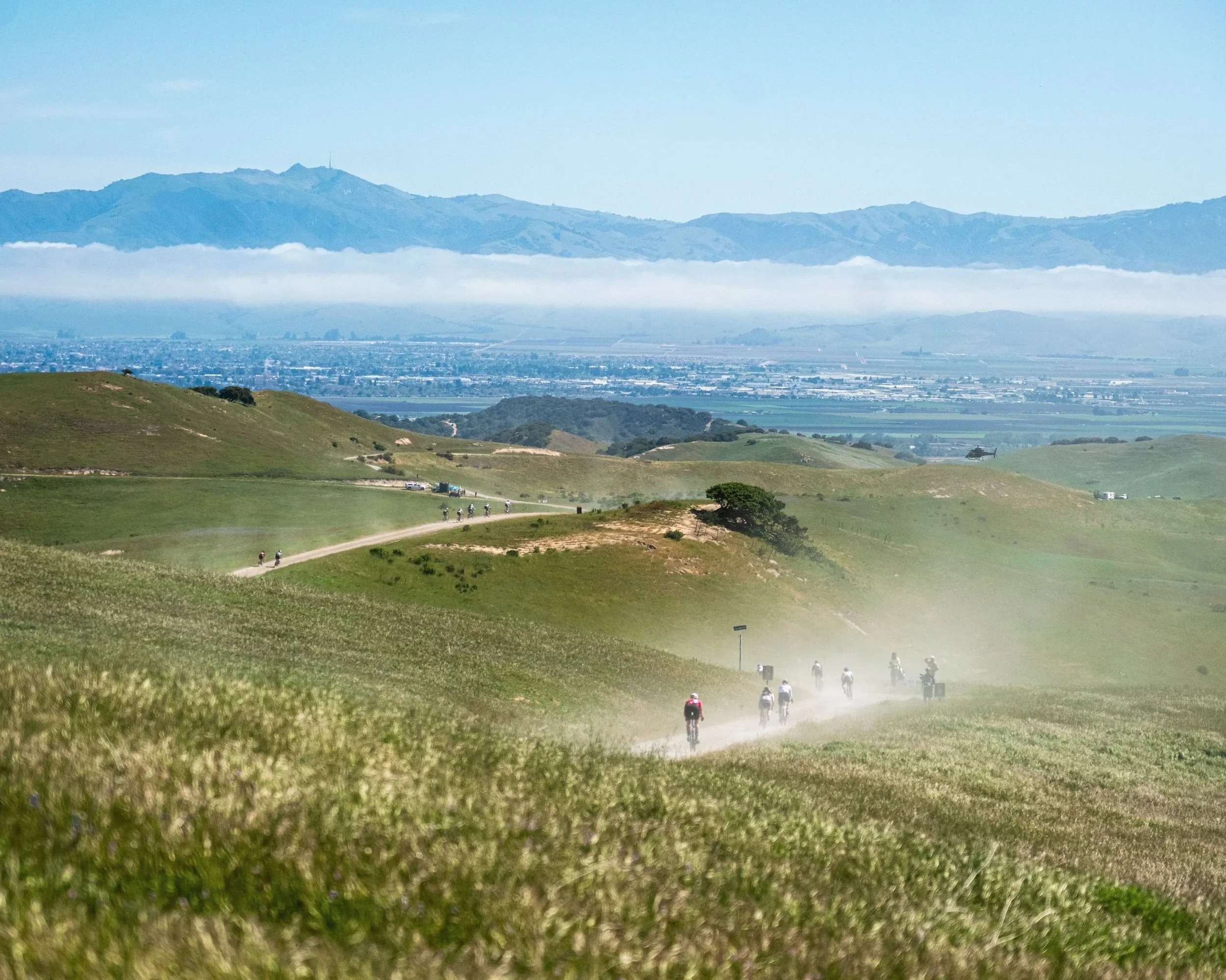 Group of cyclists riding on a dusty trail through rolling green hills with a view of mountains and a city in the distance.