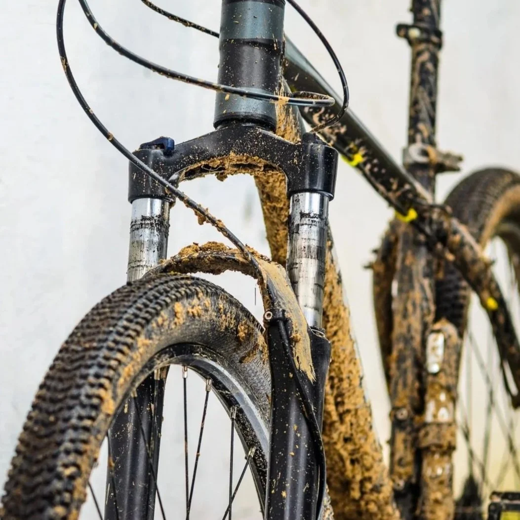 A mountain bike covered in mud, focusing on the front suspension fork and tire.