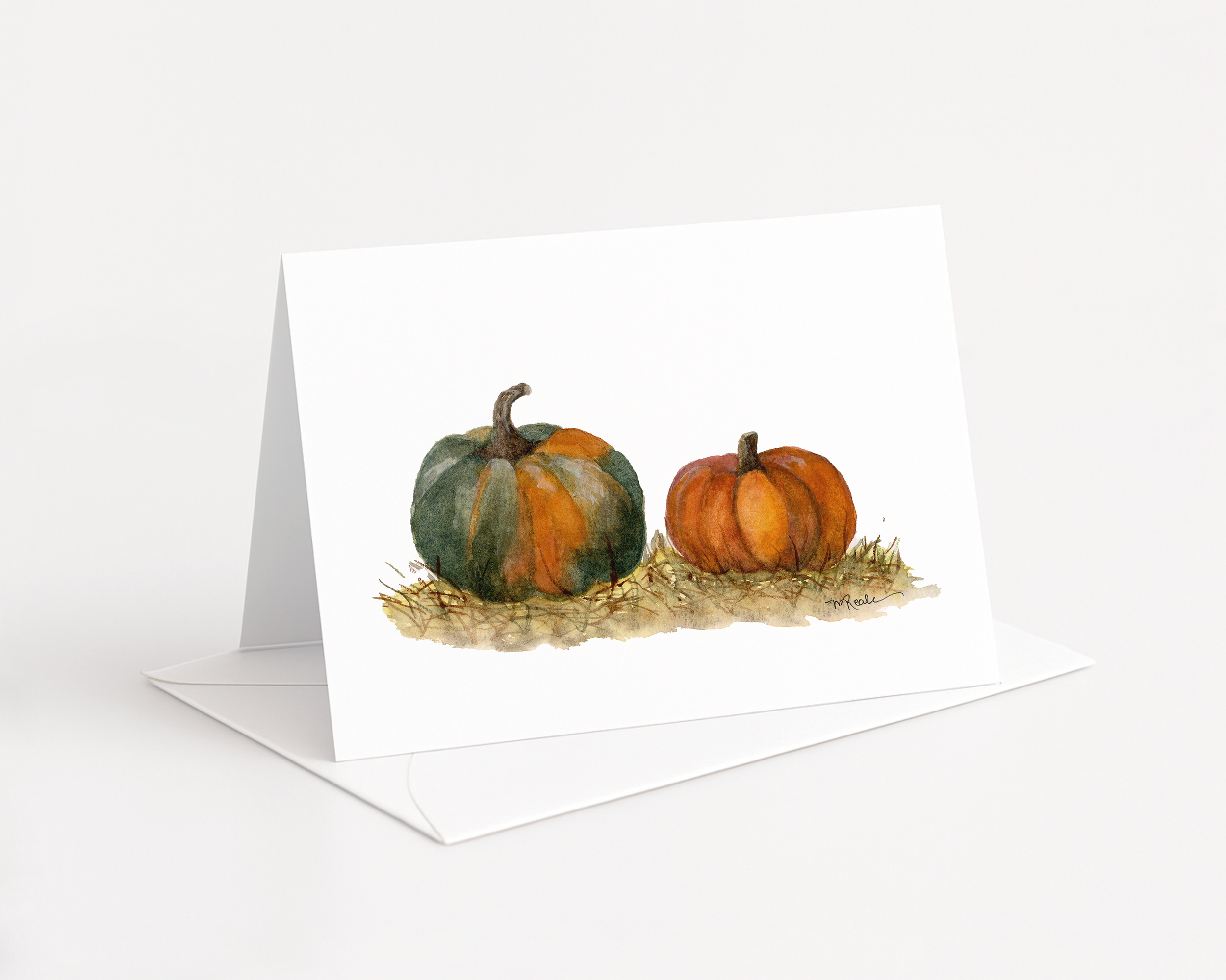 A greeting card with watercolor paintings of two pumpkins on a hay mat, placed on a white table.