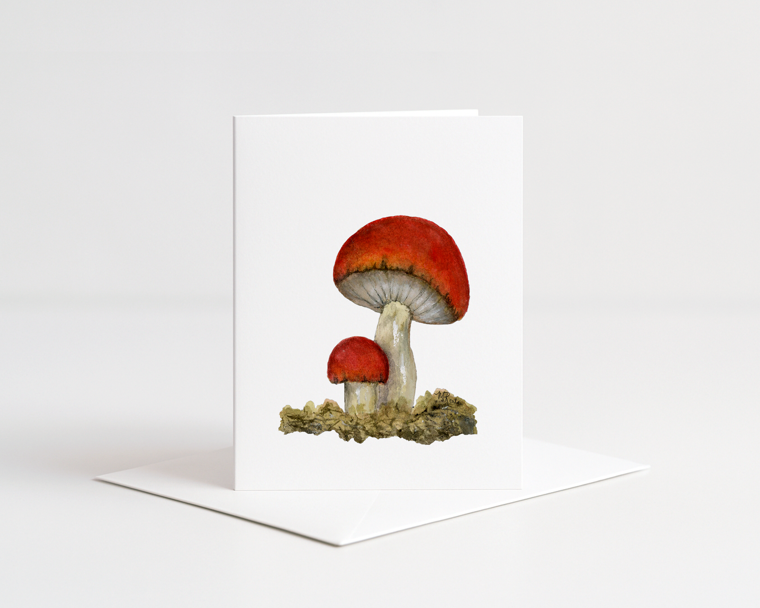 Handmade greeting card with watercolor illustration of three red and white mushrooms growing on mossy ground, signed by artist in lower right corner.
