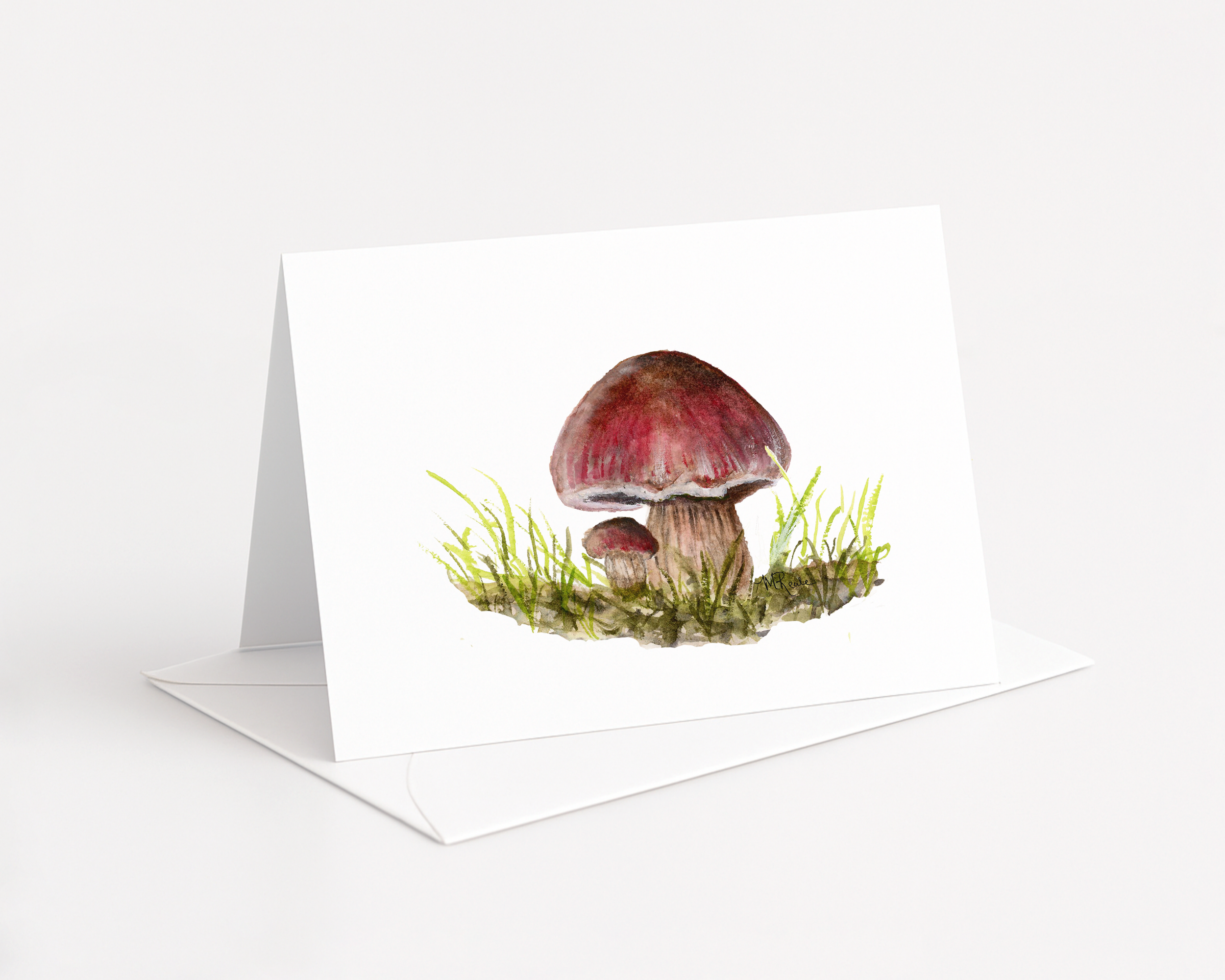 A watercolor painting of red and brown mushrooms with green grass, displayed on a white greeting card.