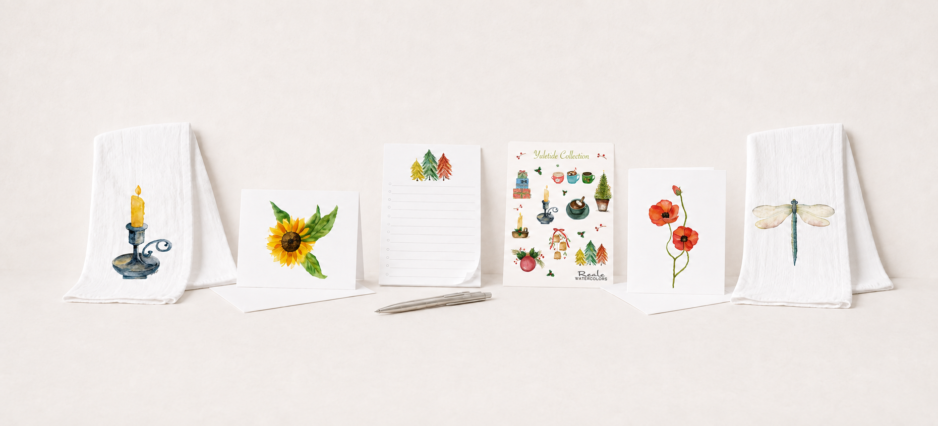 A row of greeting cards with watercolor designs, featuring a candle, sunflower, Christmas trees, holiday scenes, flowers, and a dragonfly, displayed on white napkins and a light background with a silver pen in front.