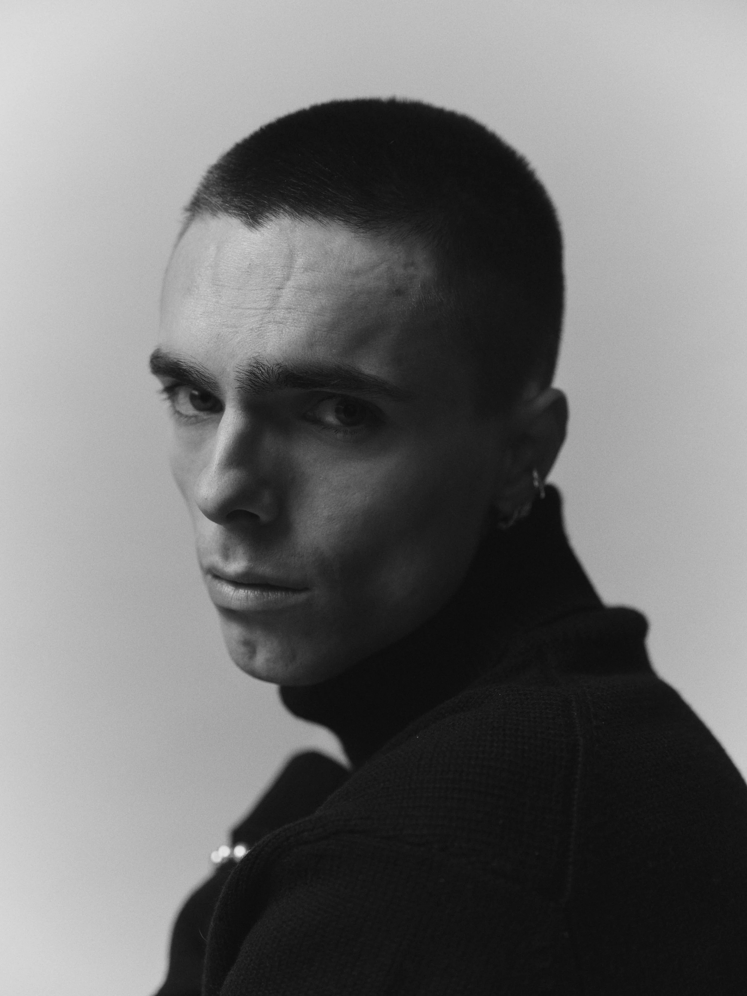 A black and white portrait of a young man with a short haircut, wearing a dark turtleneck, looking over his shoulder at the camera.