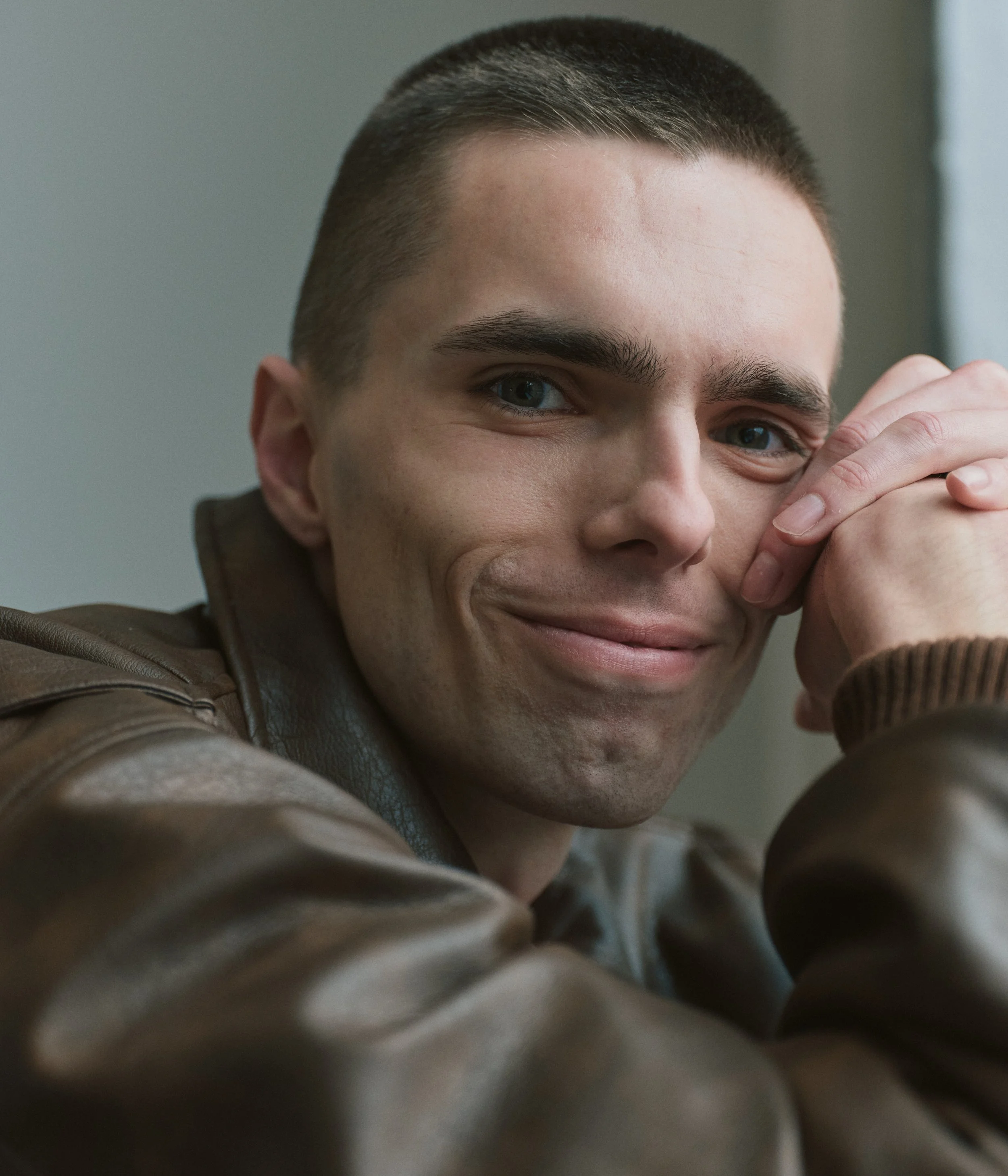 Close-up of smiling young man with short dark hair and blue eyes, wearing a brown leather jacket, resting his face on his hands.