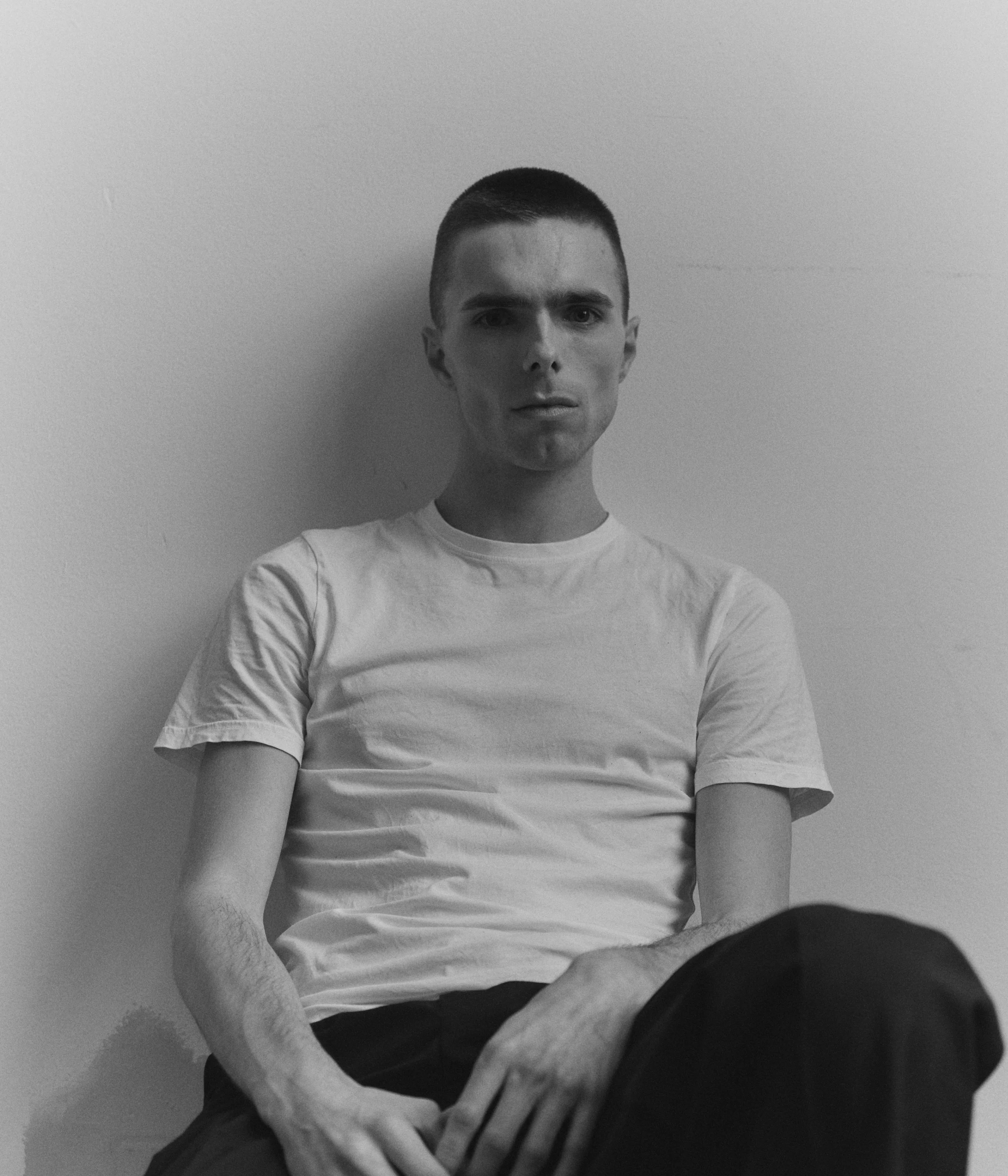 A young man with short hair sitting against a plain wall, wearing a white t-shirt and dark pants, looking directly at the camera.