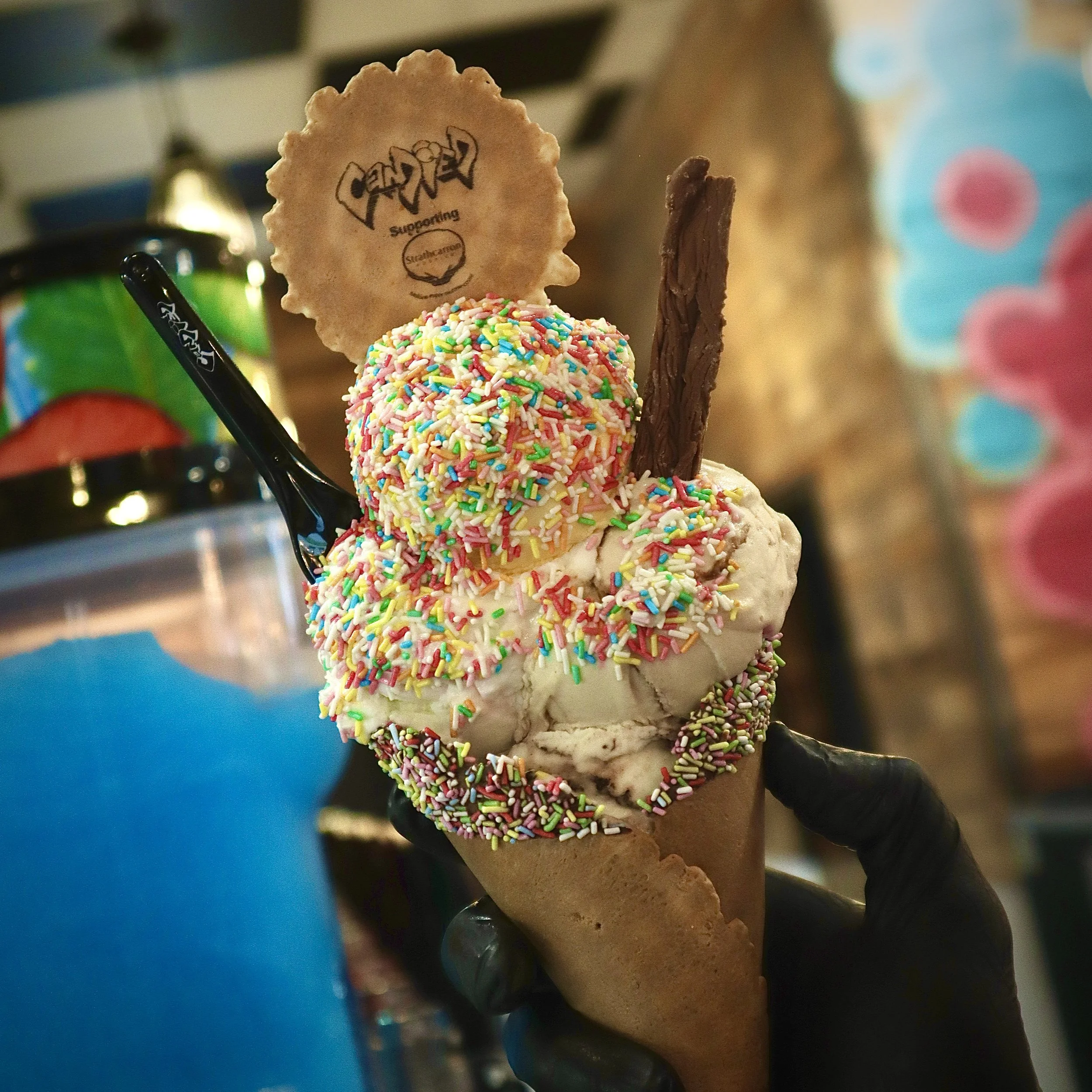 Ice cream cone with colorful sprinkles, topped with a cookie and a chocolate stick, held by a gloved hand.