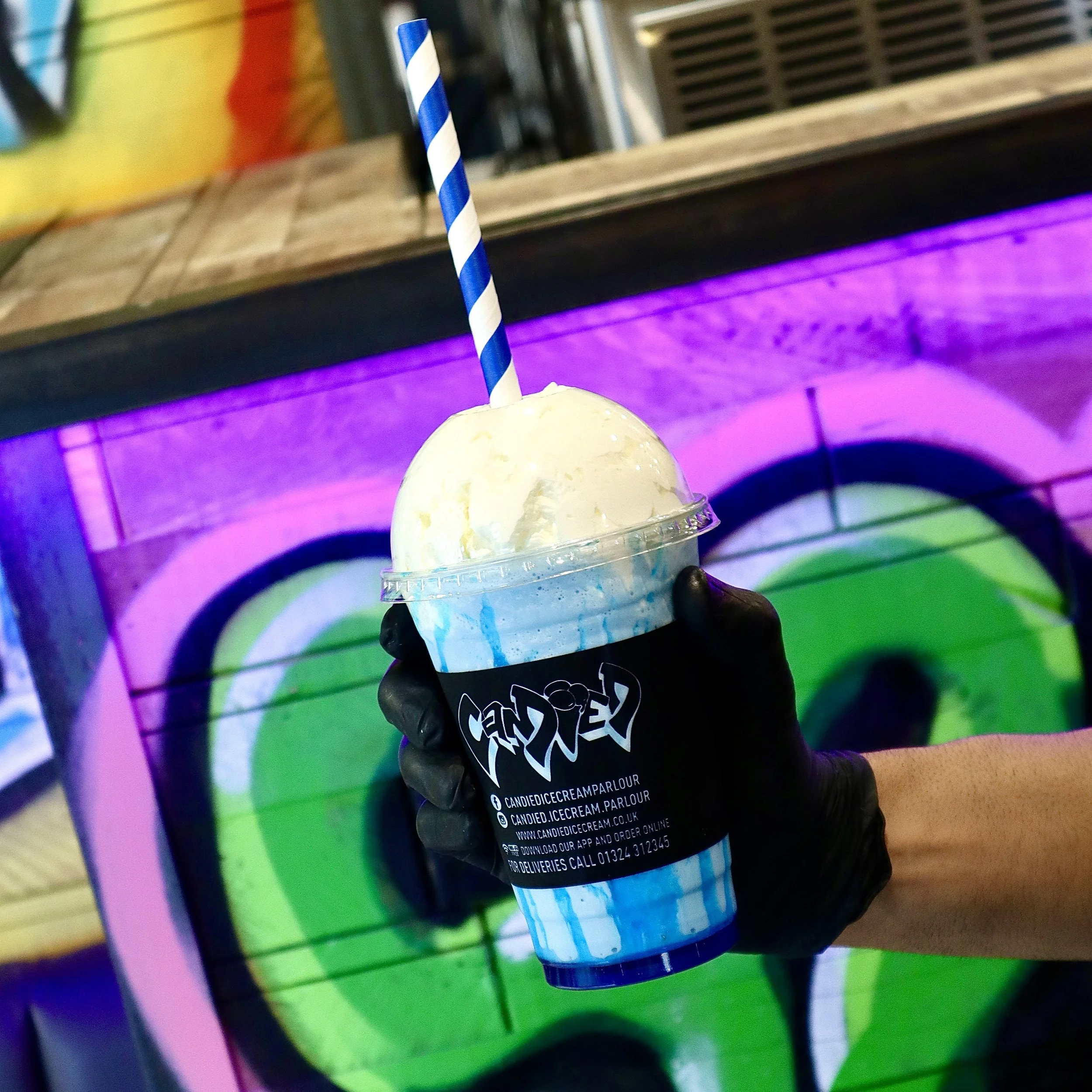 Hand wearing black glove holding a vanilla ice cream in a plastic cup with a blue and white striped straw, against a colorful graffiti wall background.