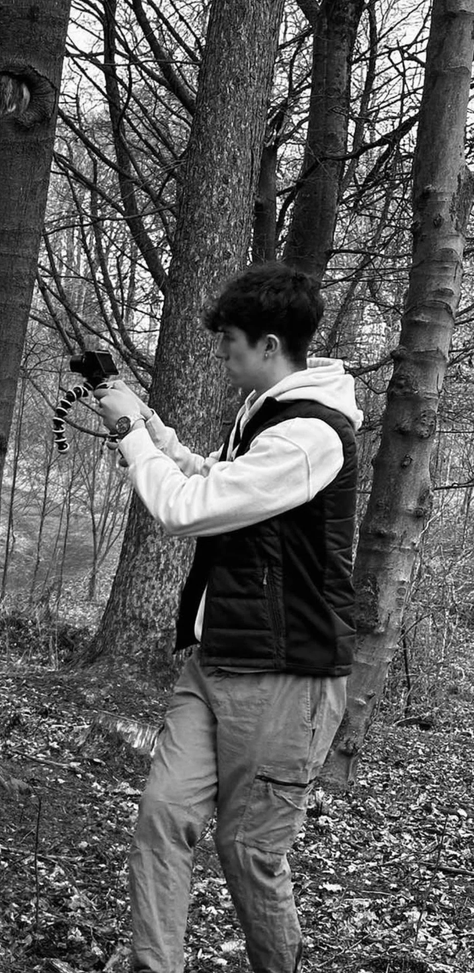 A young man in a hoodie and vest standing in a forest, holding a camera mounted on a flexible tripod, looking at the camera's screen in a black-and-white photo.