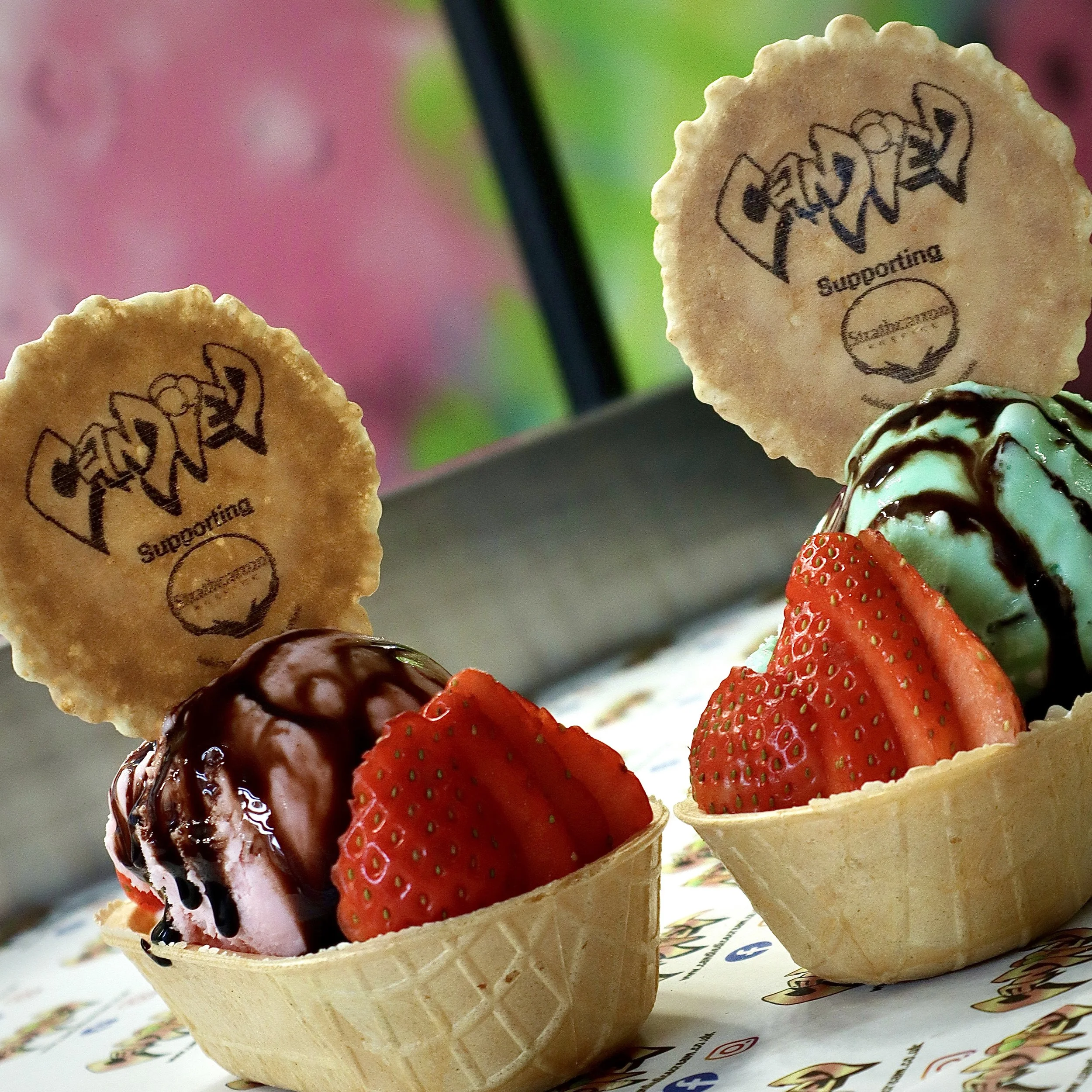 Ice cream sundaes topped with strawberries, chocolate, and mint ice cream, served in waffle bowls with supporting cookies behind them.