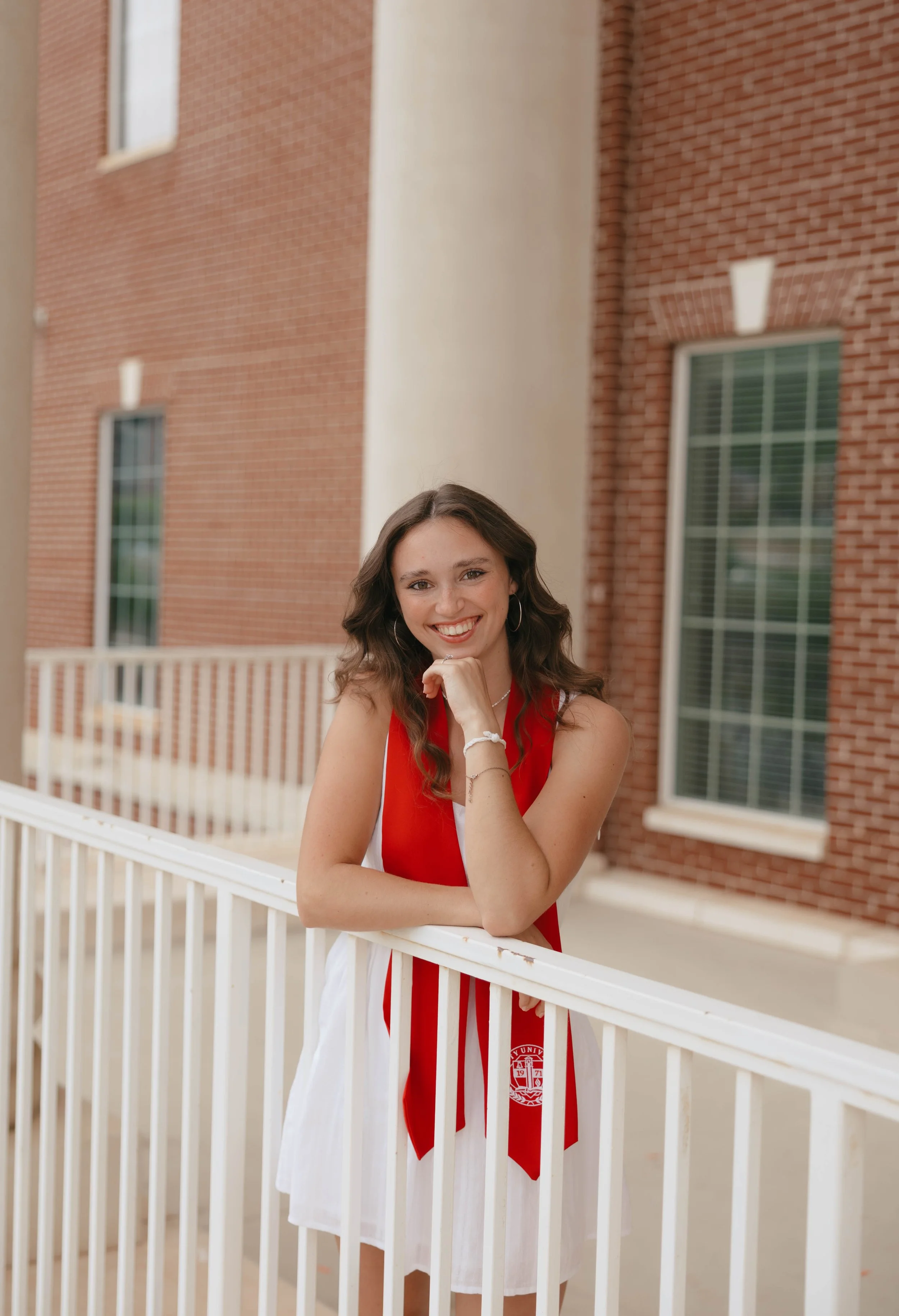 A young woman with long wavy brown hair smiling, wearing a white dress with a red stole, standing on a balcony with white railings in front of a brick building.