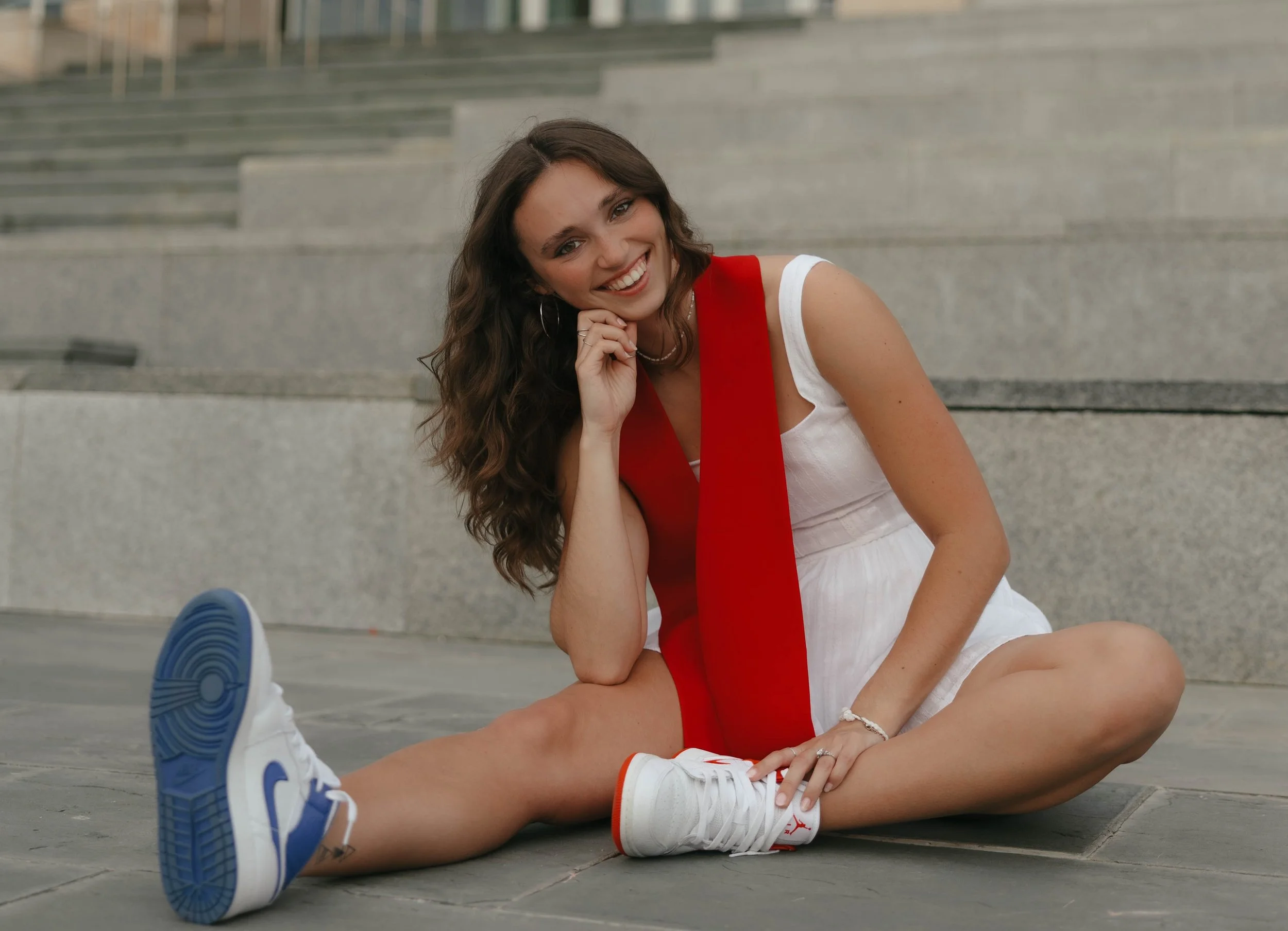 Young woman with long curly brown hair, sitting on the ground outdoors, smiling, wearing a white dress and white sneakers, with a red sash around her neck, in front of concrete stairs.