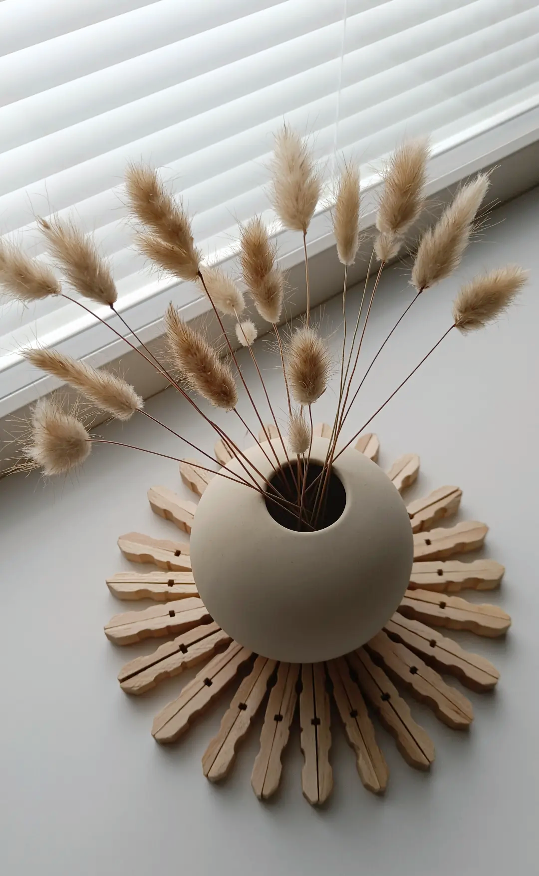 Kuva sisätiloista, jossa neutraali, pyöreä beige kukkamalja ja siinä kuivakukkia, asettuna tasolle, edessä valkoinen ikkunalauta ja ikkunassa valkoiset sälekaihtimet.