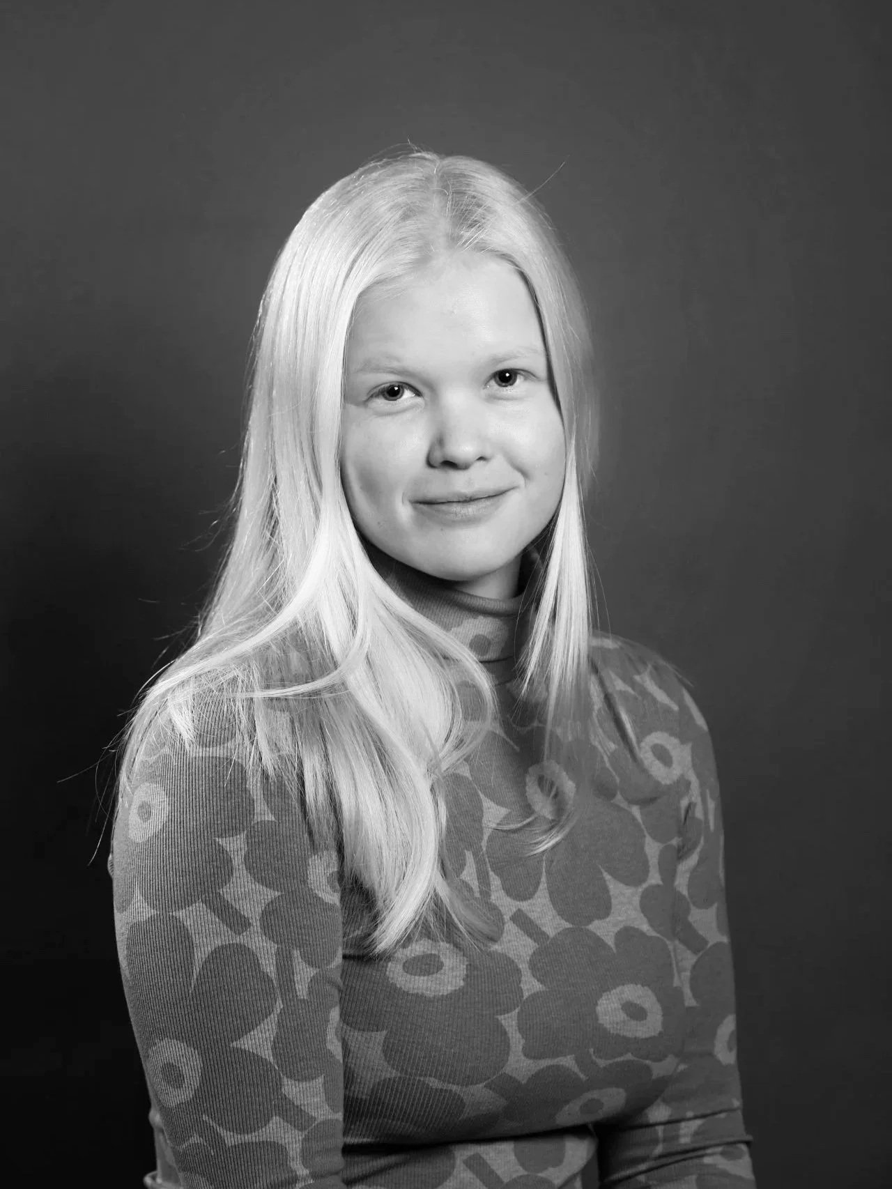 Mustavalkoinen valokuva nuoresta naisesta, jolla on pitkät suorat hiukset ja lämmin hymy, pukeutuneena ruutukuosiseen puseroon, taustalla yksinkertainen harmaa seinä.