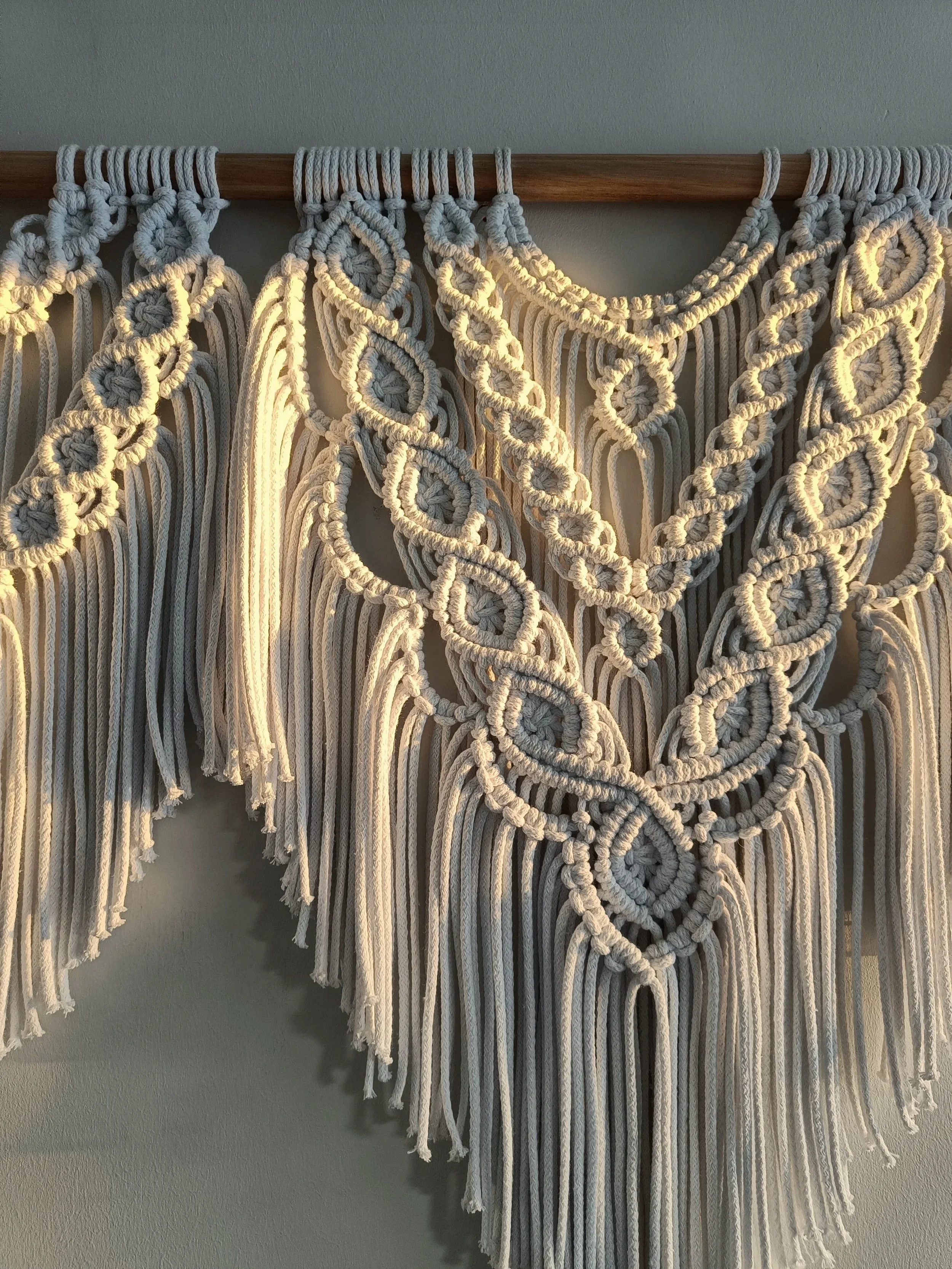 Käsityömatto tehty makramee-tekniikalla, ripustettuna puukoukkuun, luonnonväristä lankaa.