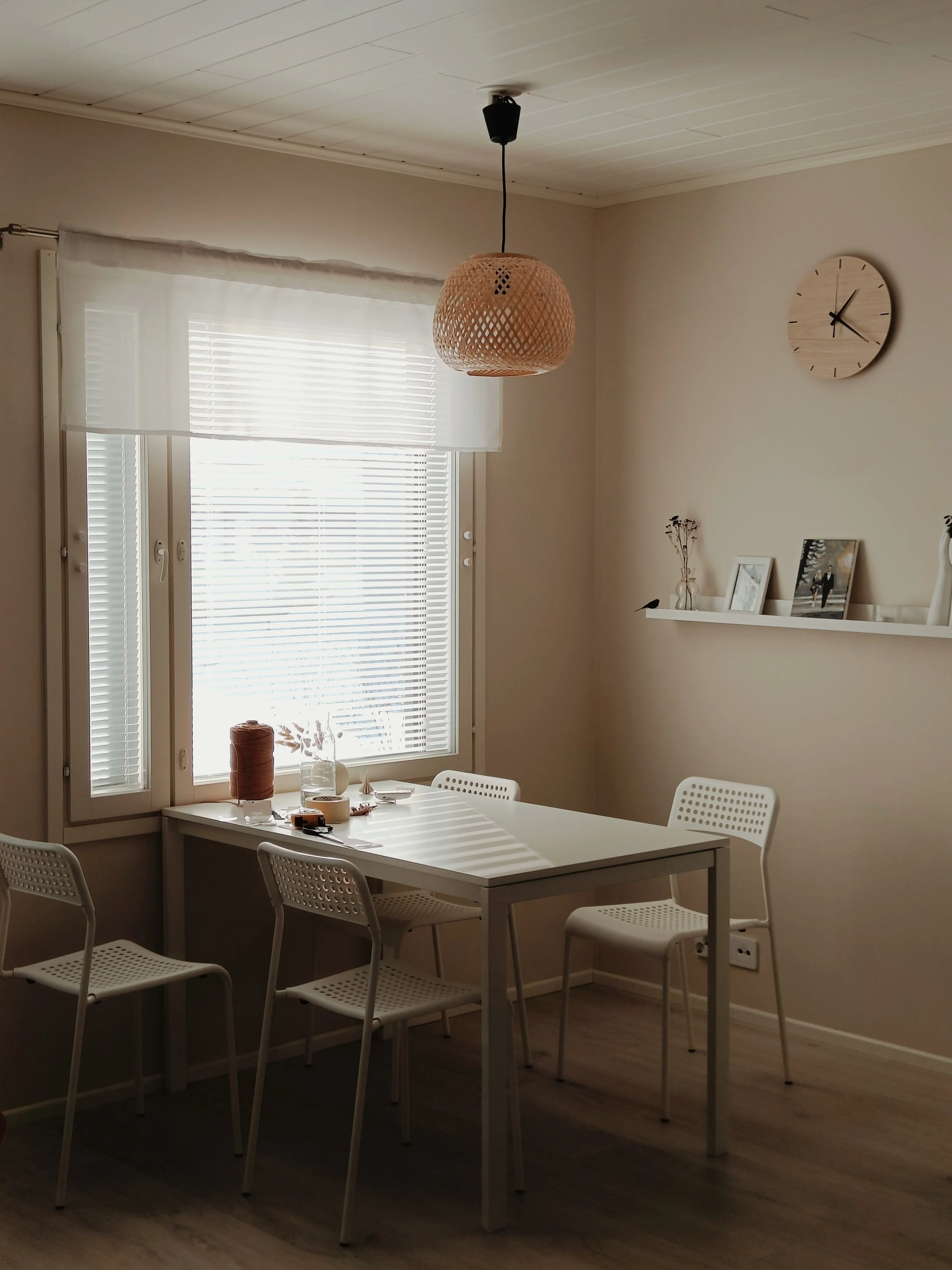 Kuva modernista kodin ruokailutilasta, jossa on valkoinen pöytä ja neljä valkoista muovituolia. Ikkunassa on valkoiset kaihtimet ja verhot. Katossa roikkuu rautainen kattovalaisin. Seinällä on ympyrän muotoinen puinen kello ja seinähyllyllä valokuvia ja koriste-esineitä.