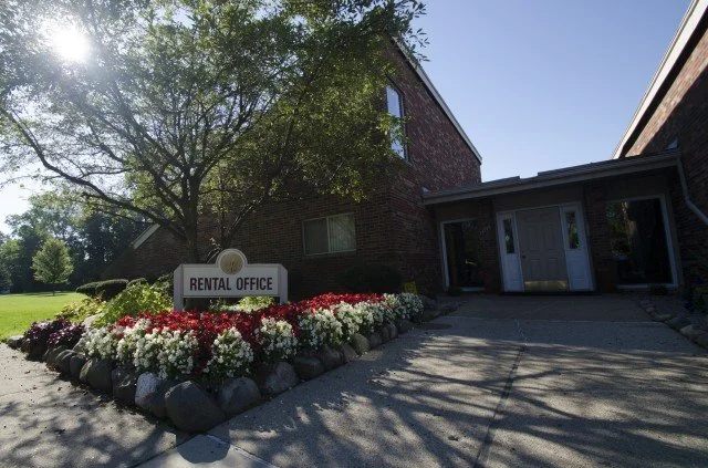 Exterior view of a rental office building with a sign in front, surrounded by a flower bed with red and white flowers, and a large tree partially shading the building.
