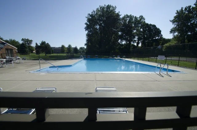 Empty outdoor swimming pool with lounge chairs and a fence surrounding it, in a sunny setting with trees in the background.