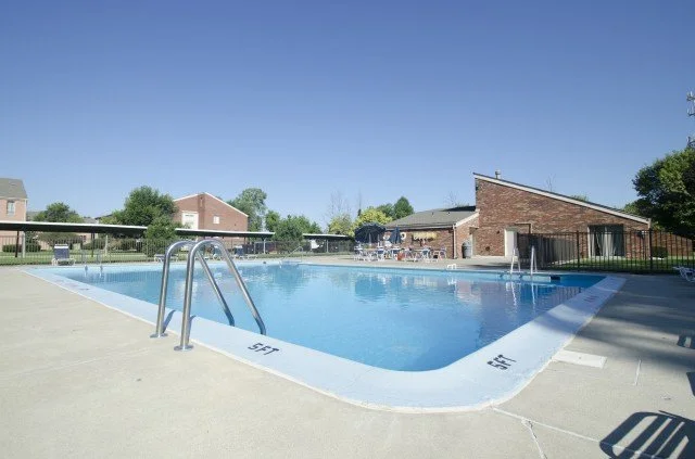 Empty outdoor swimming pool with metal handrails, surrounded by a concrete deck, with residential buildings and green trees in the background under a clear blue sky.