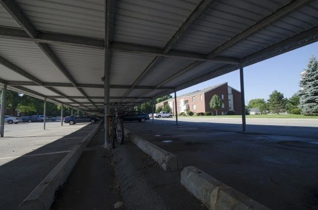 Empty parking lot partially covered by a metal roof, with a bicycle leaning against a concrete barrier in the foreground, and an apartment building and trees in the background.