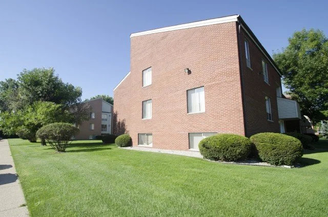 Apartment building with brick exterior, surrounded by a well-maintained lawn and trimmed bushes under a clear blue sky.