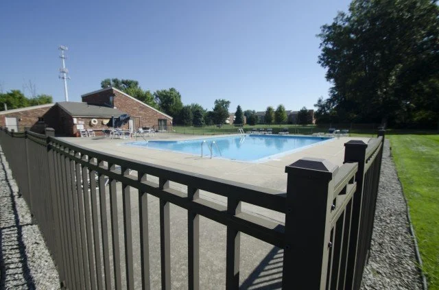 Outdoor swimming pool surrounded by a black metal fence, with a brick building and trees in the background.