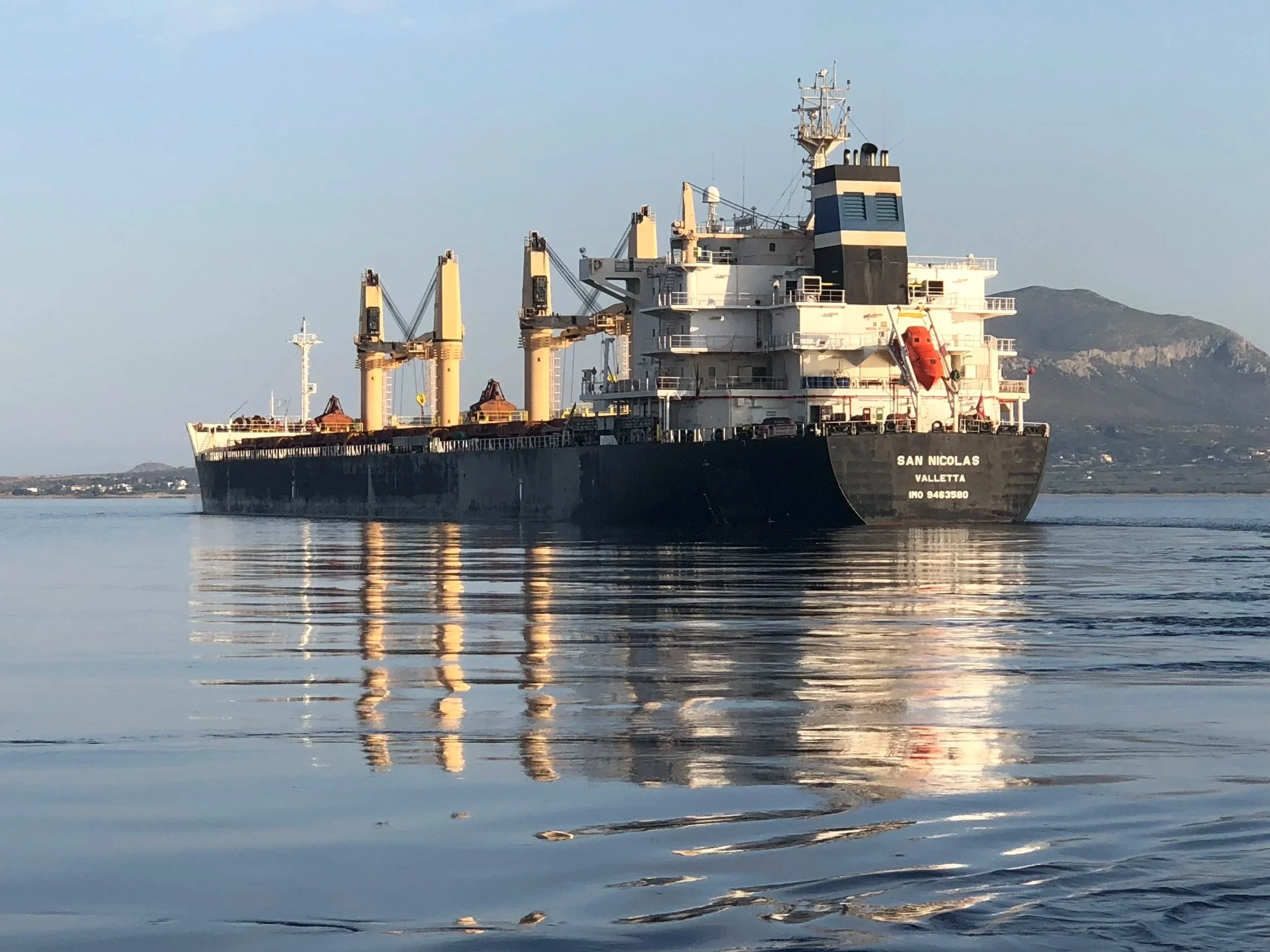 Large black cargo ship named San Nicolas with white superstructure and four cranes, sailing in calm water with a mountainous landscape in the background.