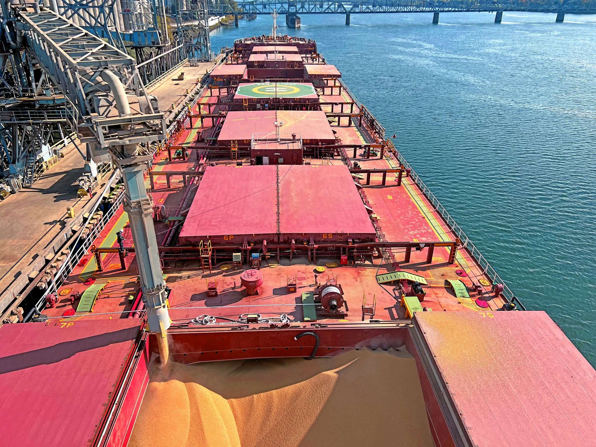 A large cargo ship is unloading a yellow cargo onto the water at a port, with a bridge visible in the background.
