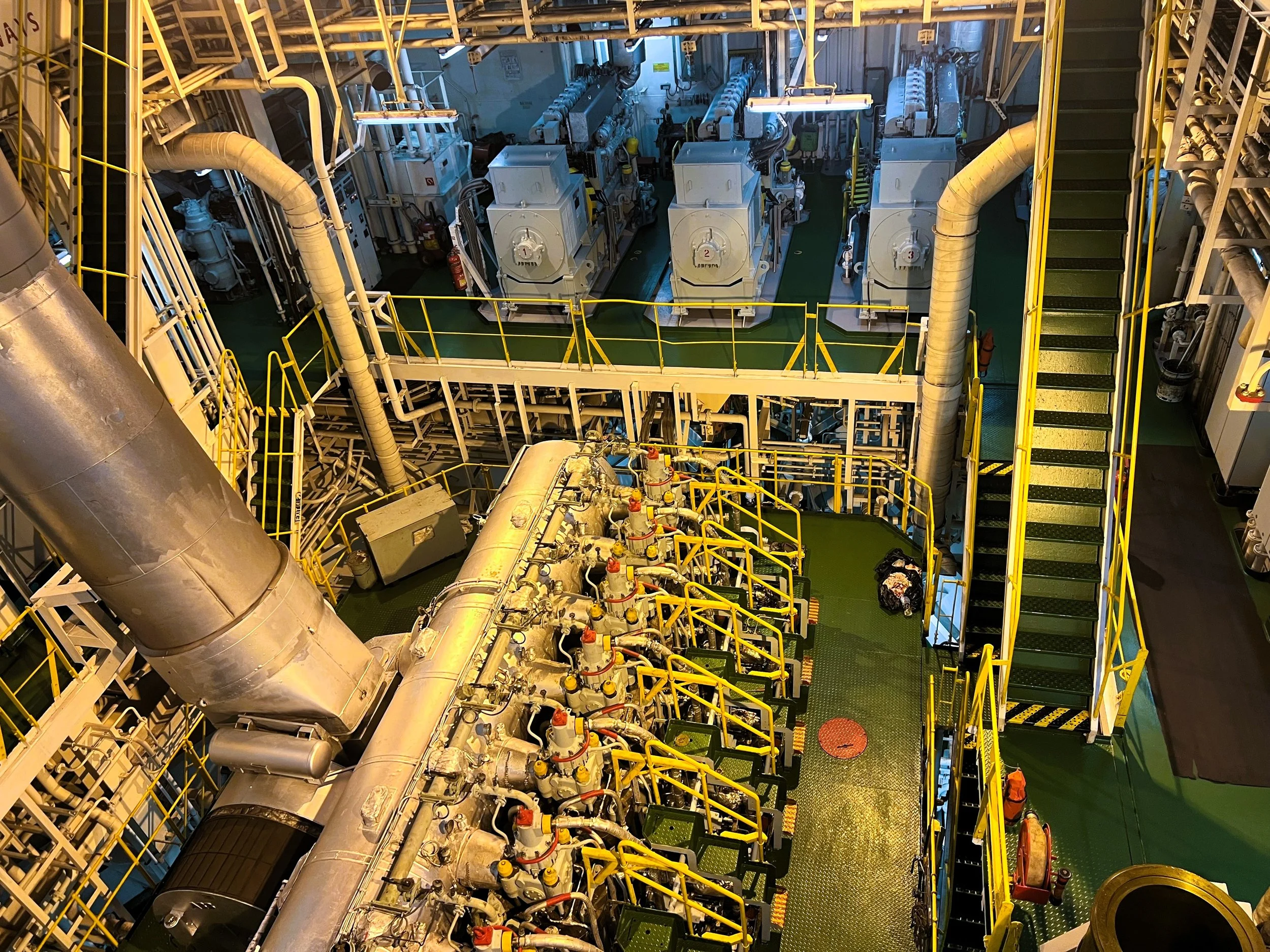 Engine room with large pipes, machinery, and yellow safety railings, seen from above.