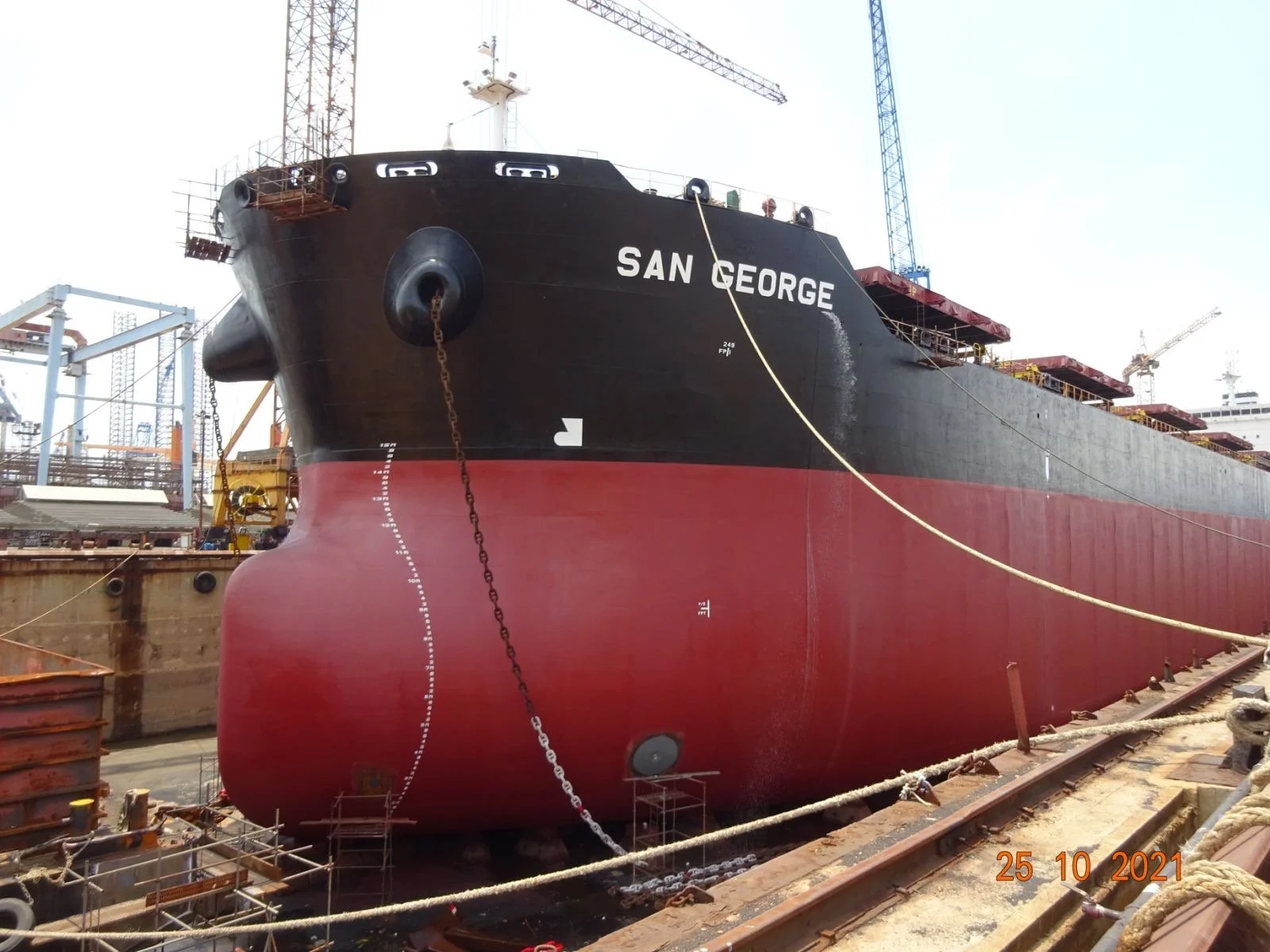 Close-up of the bow of a large black and red bulk carrier ship named 'San George' docked at a shipyard with cranes in the background.