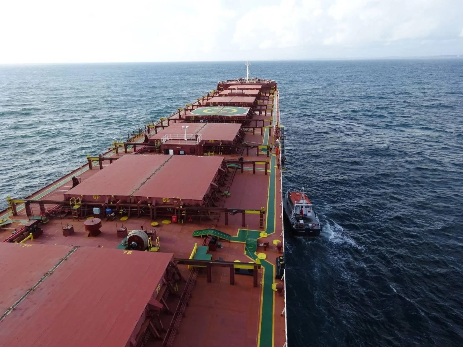 View from above of a large red cargo ship sailing on the ocean, with a small tugboat guiding alongside.