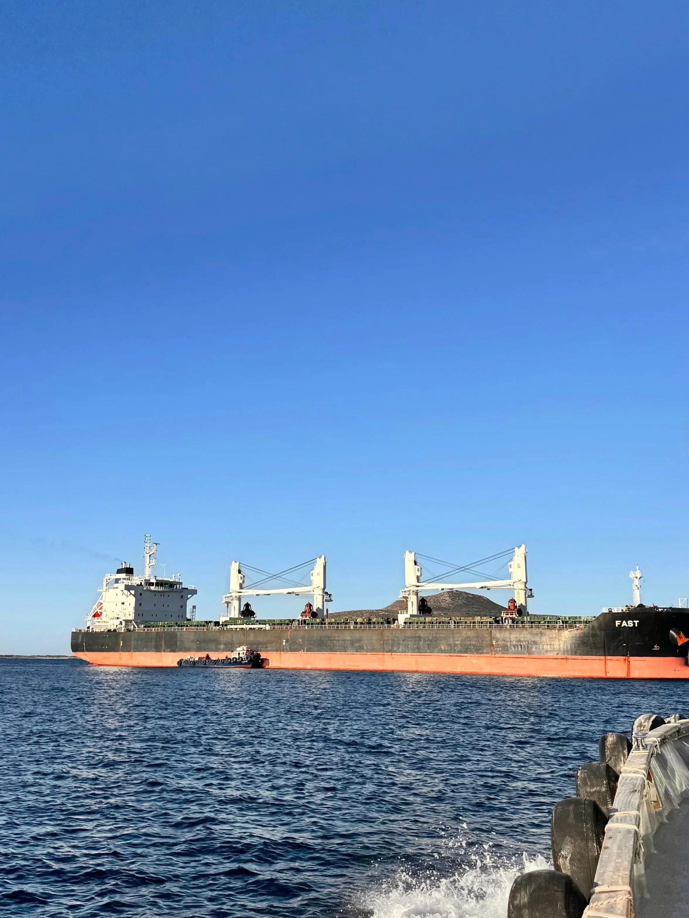 A large cargo ship named 'FAST' sailing on the ocean beside a dock with large rubber bumpers.