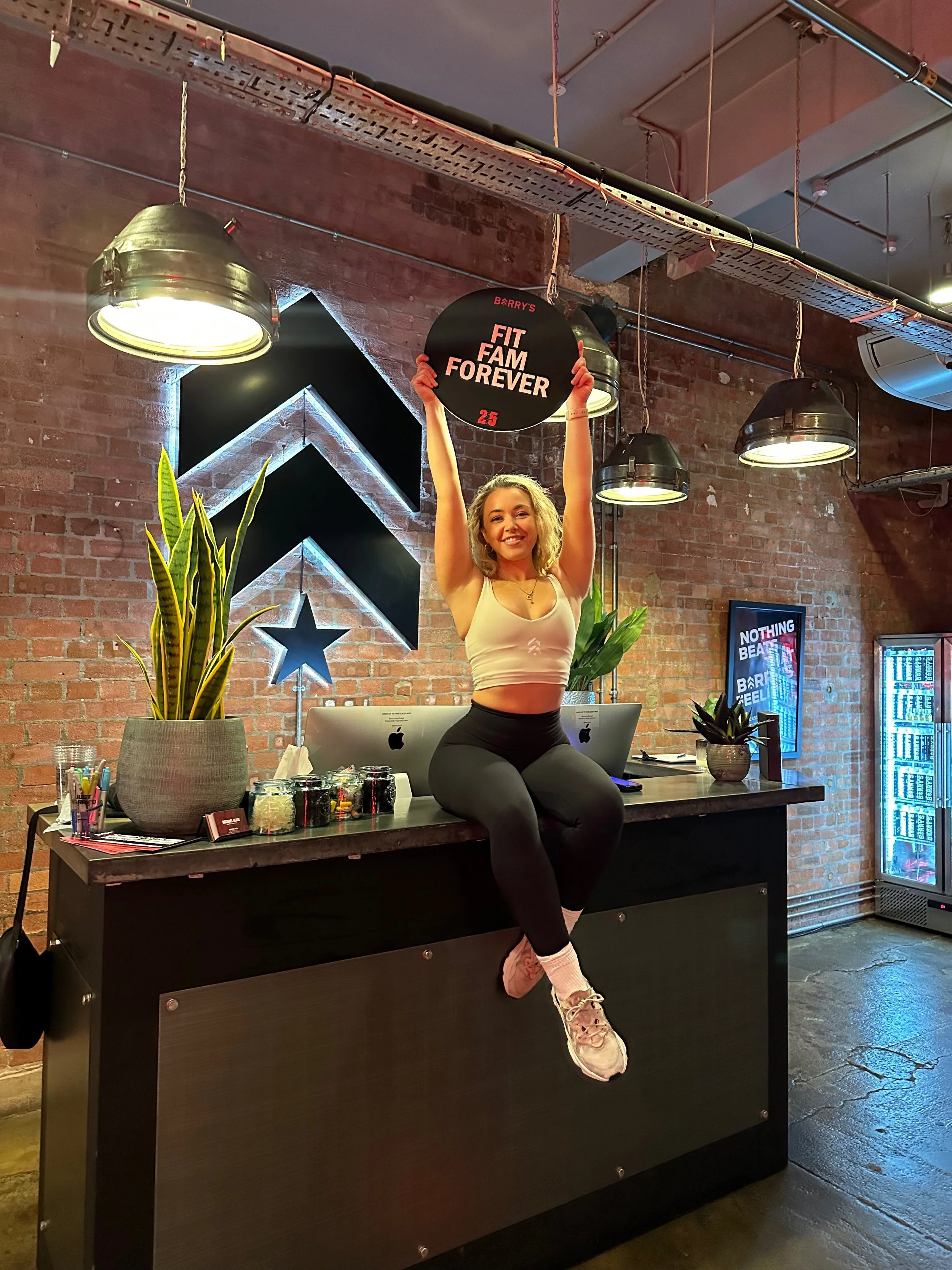 A woman sitting on a counter in a gym, holding a black circle sign that says 'Barry's Fit Fam Forever' with the number 25. She is wearing a white sports bra, black leggings, pink sneakers, and has blonde curly hair. There are plants, computers, and fitness-related decor in the background.
