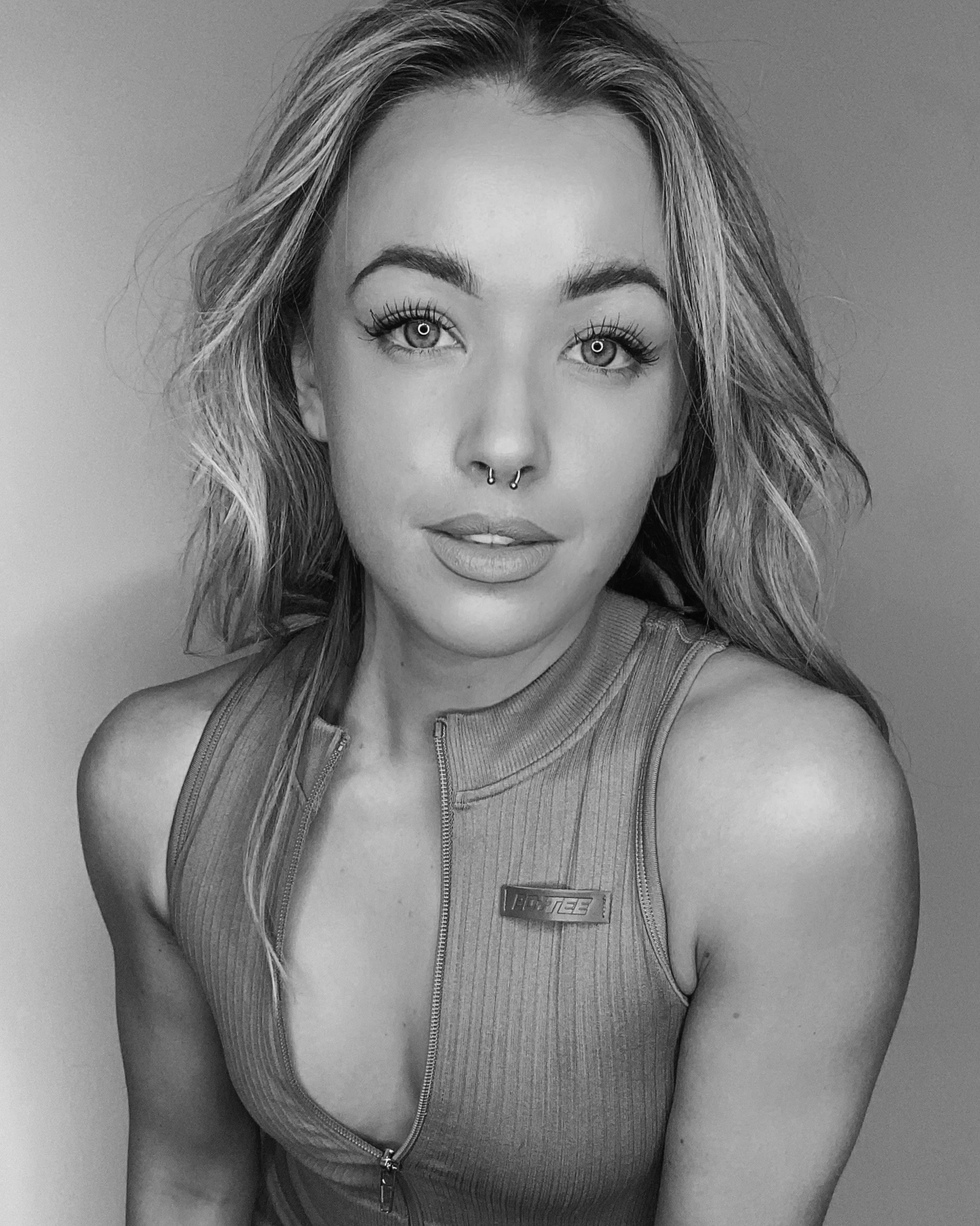 Black and white portrait of a woman with wavy hair, wearing a sleeveless top with a zipper, looking directly at the camera.