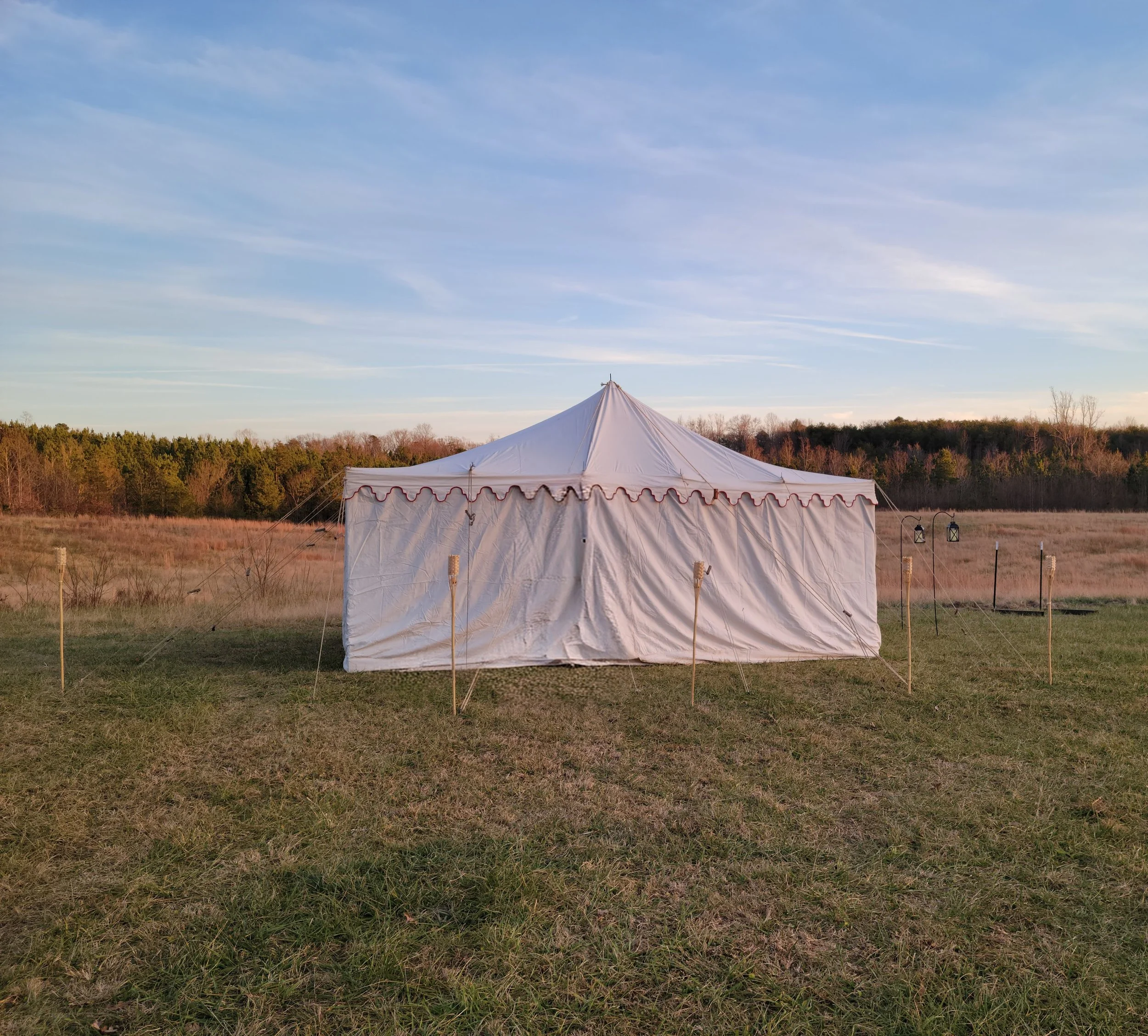 A large white event tent set up outdoors in a grassy field with a backdrop of trees and a blue sky.