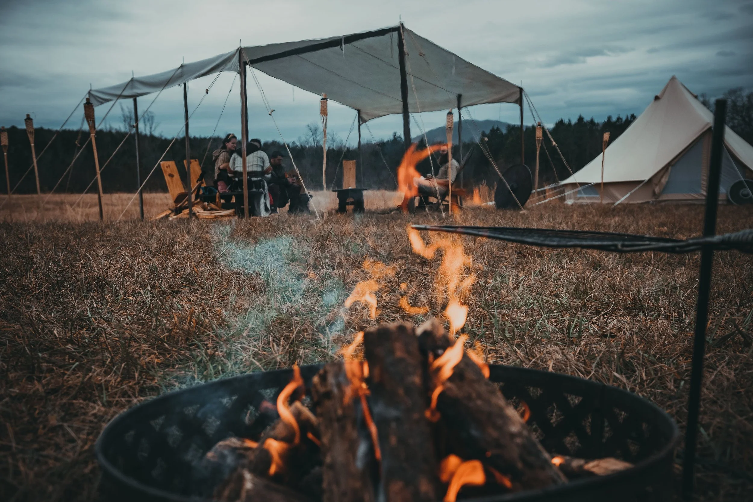 A campfire with burning logs in the foreground, with a group of people sitting under a large outdoor canopy and tents in the background on a cloudy day.