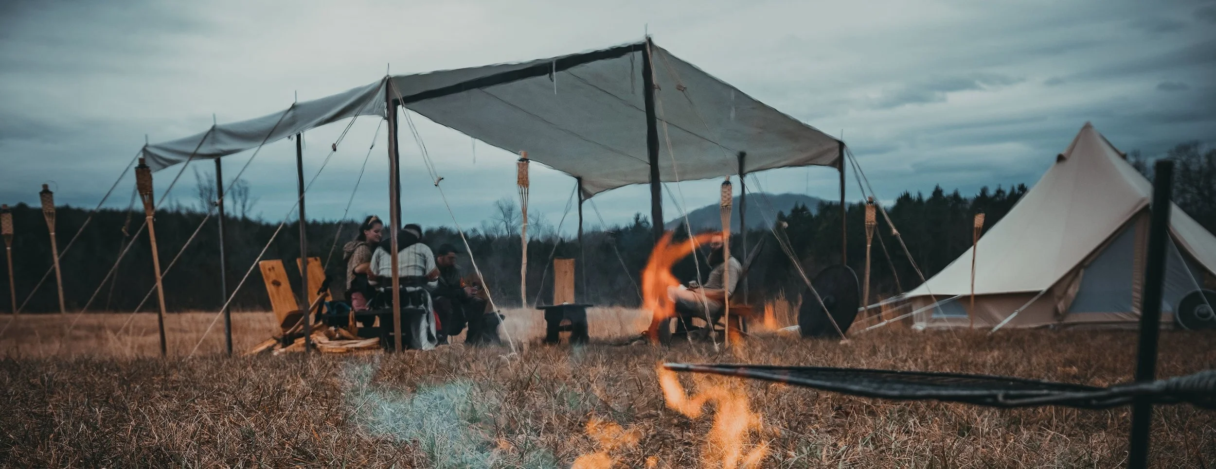 Group of people sitting under a tent near a campfire in an open field with tents and a forested area in the background.