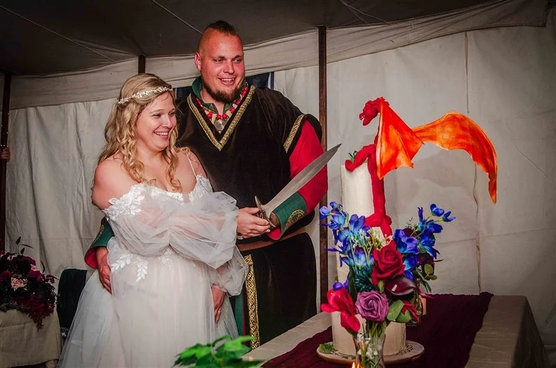 A bride and groom in medieval costumes cutting a wedding cake decorated with a red dragon and purple and red flowers.