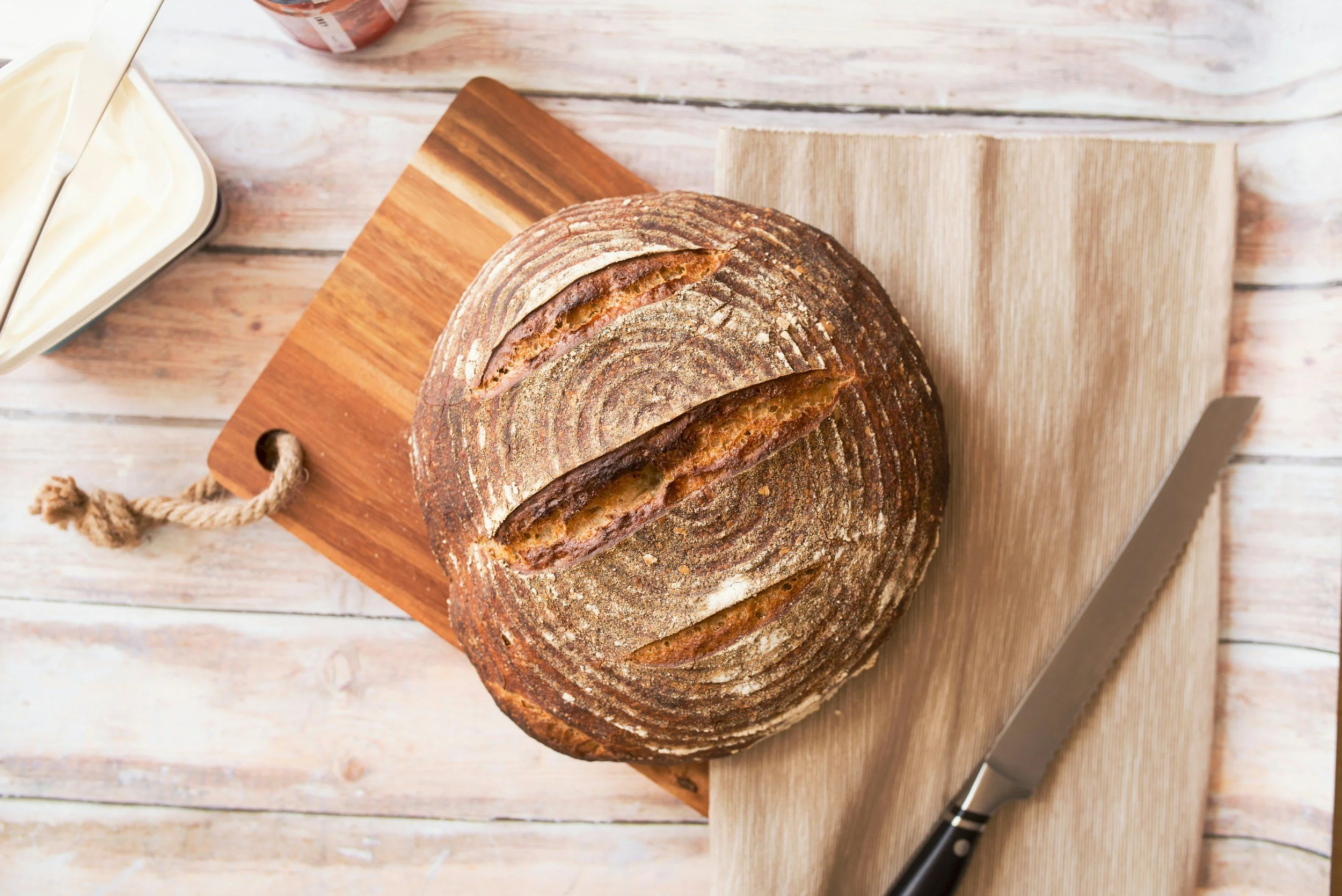 Round sourdough bread on a wooden cutting board with a serrated knife and a butter dish nearby.
