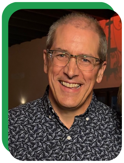 A smiling middle-aged man with glasses, wearing a patterned shirt, in an indoor setting.