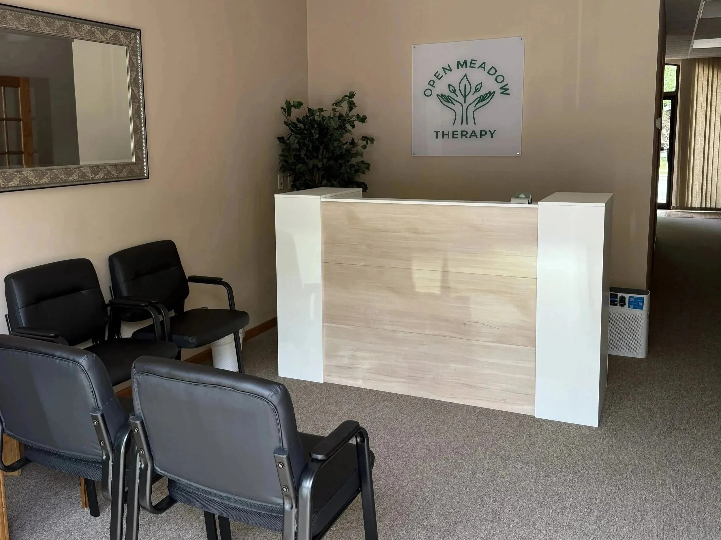 Reception area at Open Meadow Therapy with black chairs arranged in a row, a white reception desk, a green leafy plant, a mirror on the wall, and a sign that reads 'Open Meadow Therapy' with a tree logo.
