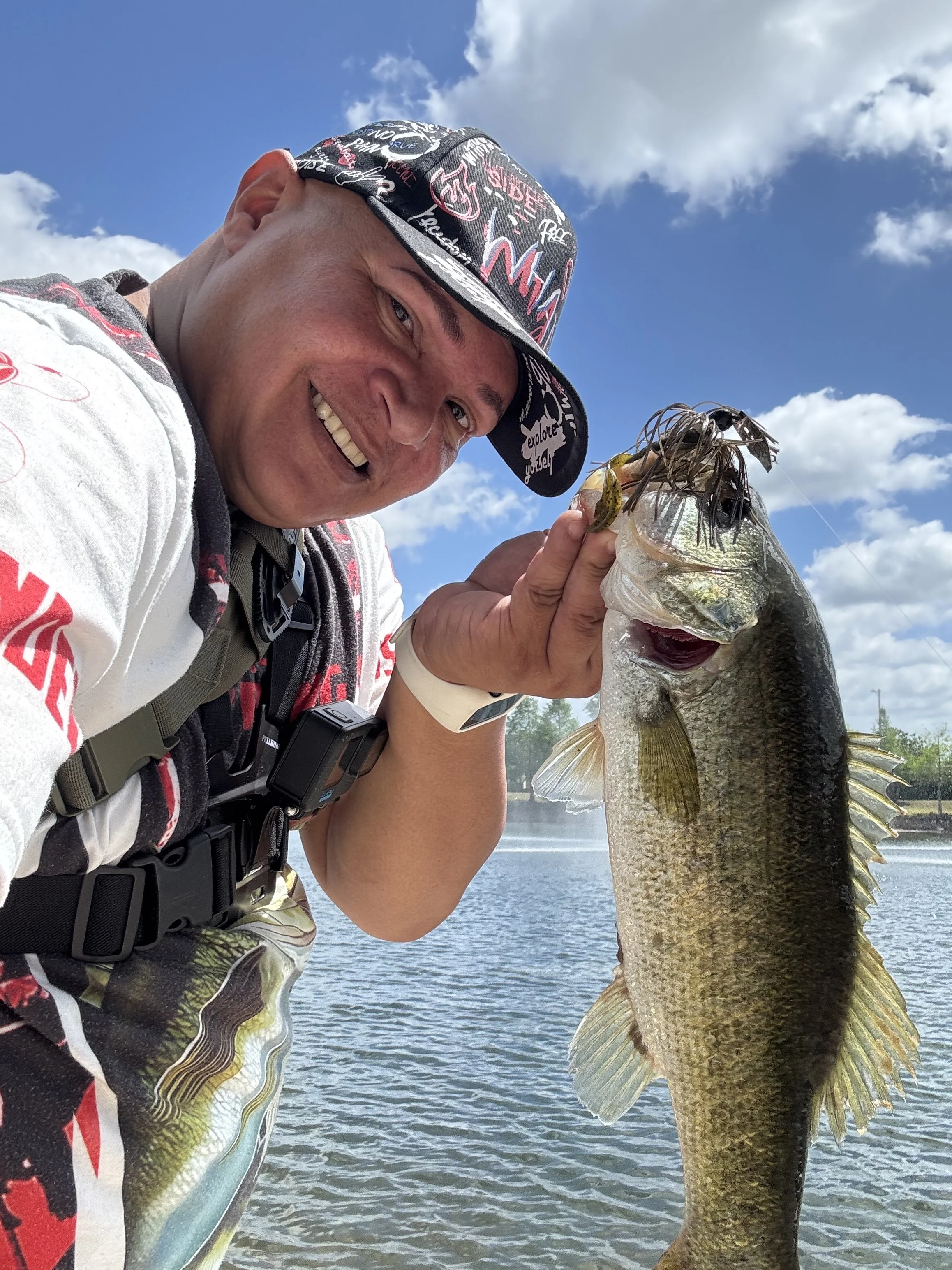 Man smiling and holding a large bass fish he caught, outdoors near a body of water, with blue sky and clouds in the background.
