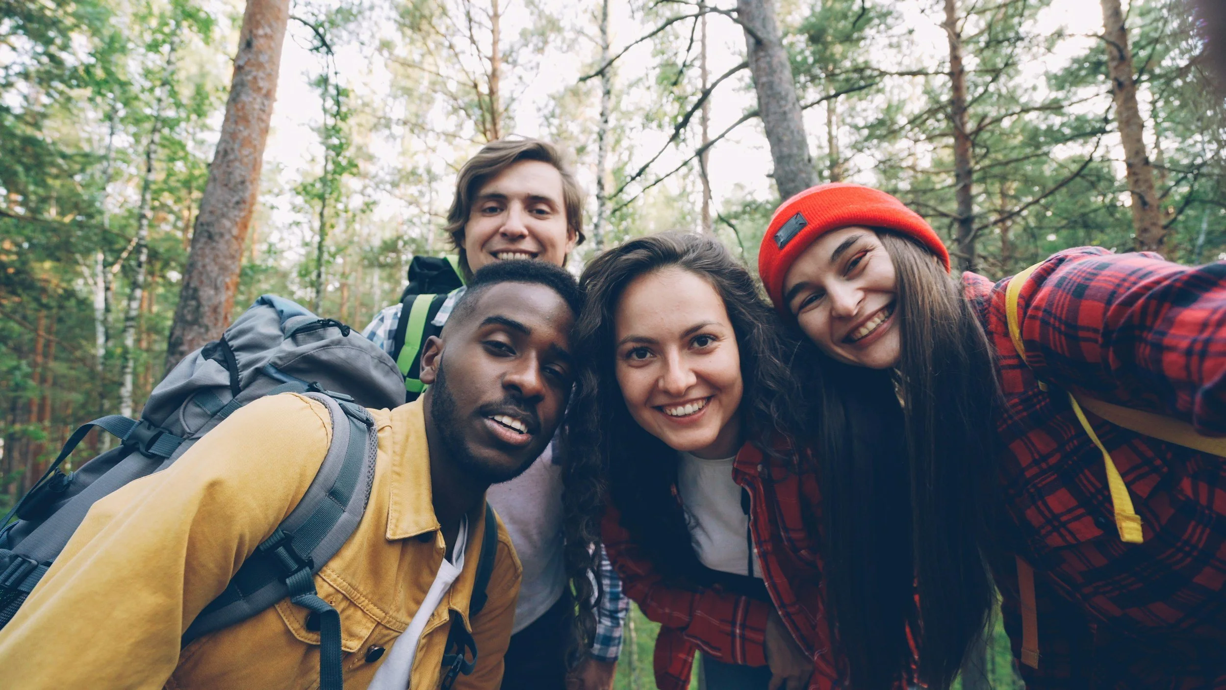 Group of five young friends taking a selfie in a forest, smiling and dressed casually with backpacks and outdoor clothing.