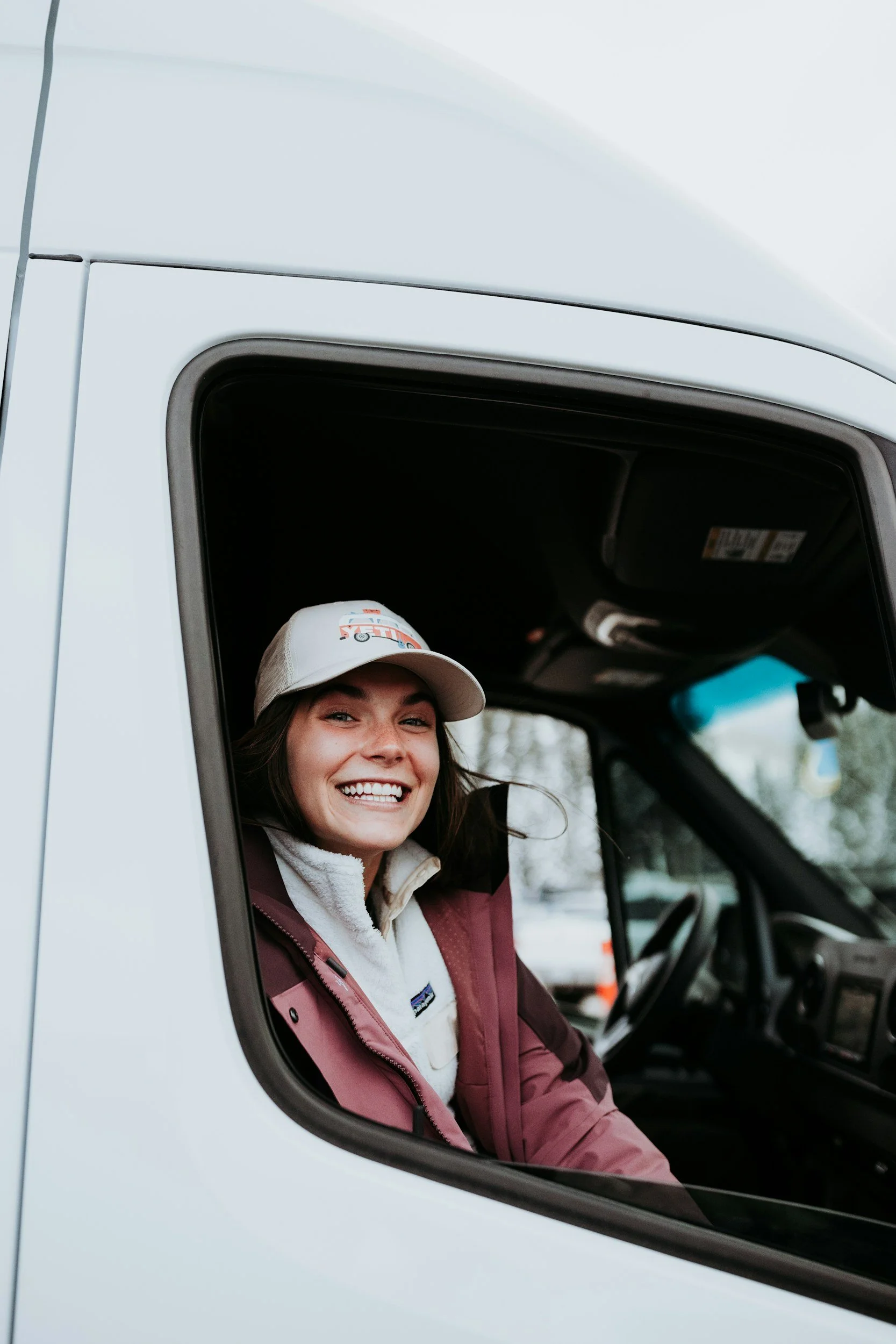 Young woman smiling while sitting in the driver's seat of a white vehicle, wearing a baseball cap and outdoor clothing.