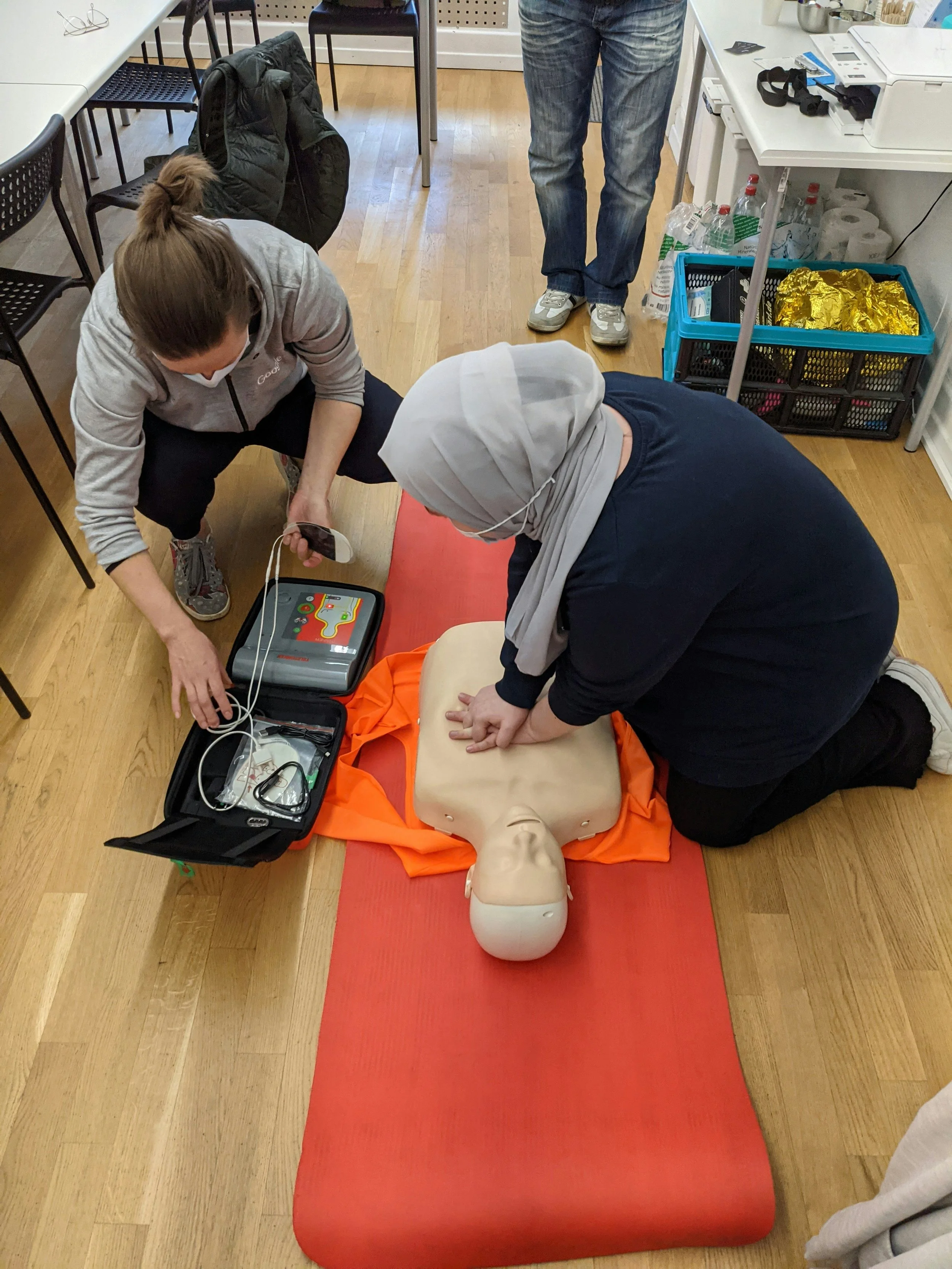 A medical training session showing two people performing CPR on a mannequin on a red mat. One person is kneeling and the other is lying beside the mannequin. There is a first aid kit, medical equipment, and supplies in the background.