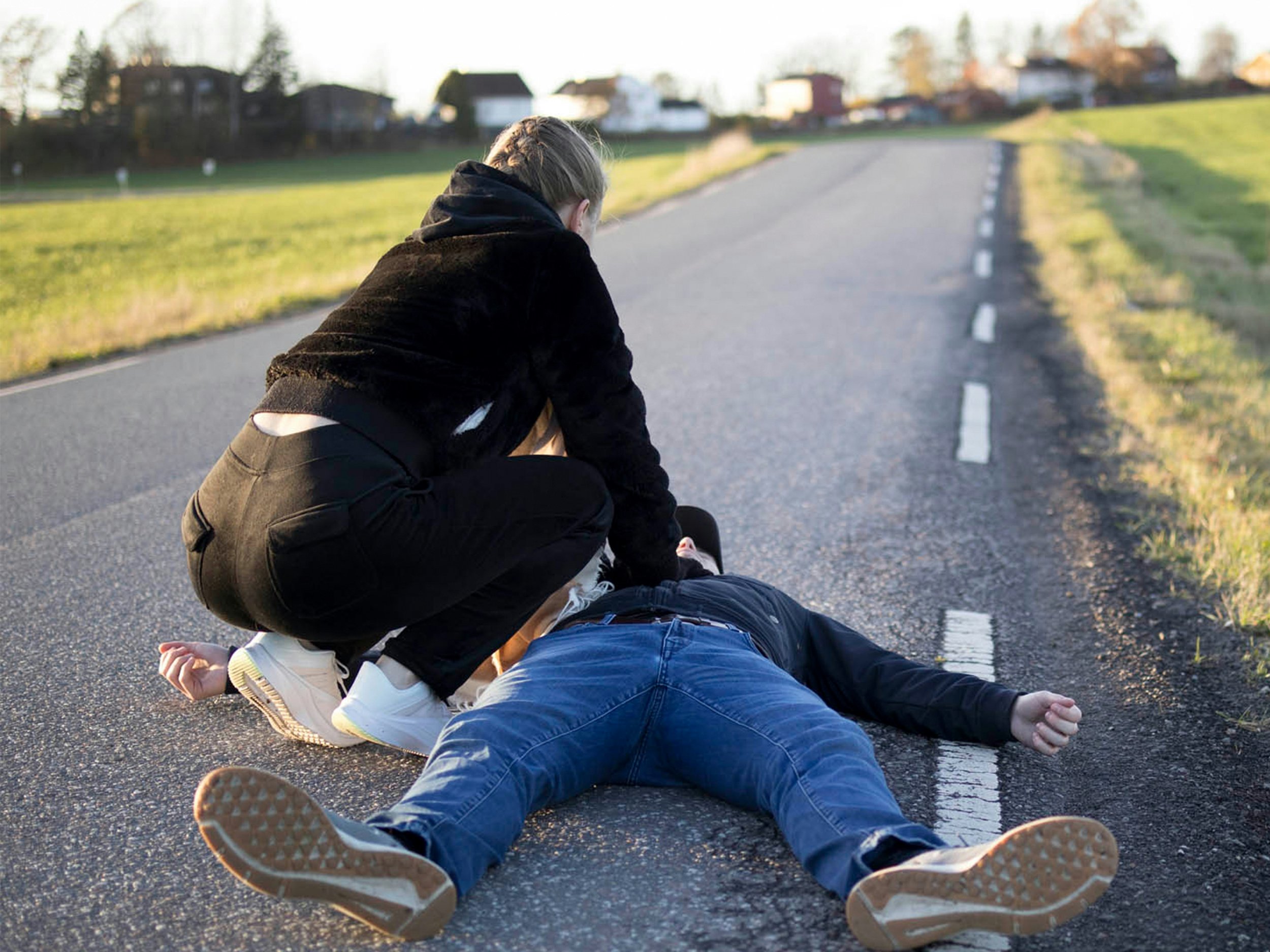 Person lying on the ground after falling on a road, with another person crouching nearby, suggesting a possible accident or medical emergency.