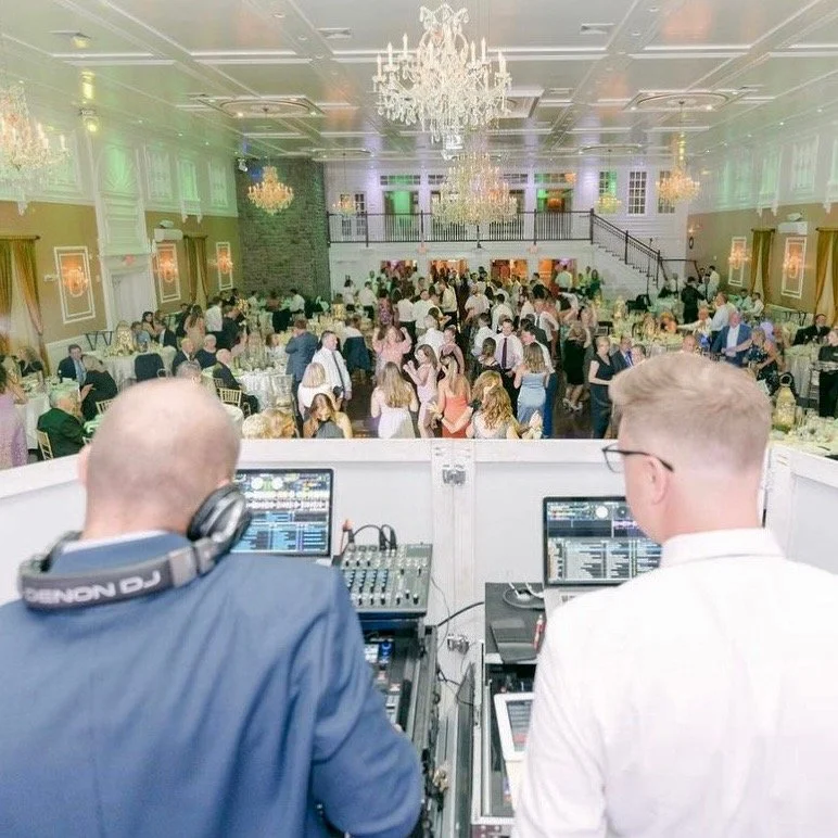 Two DJs are playing music at a reception hall filled with dancing guests, chandeliers hanging from the ceiling, and a staircase leading to an upper level.