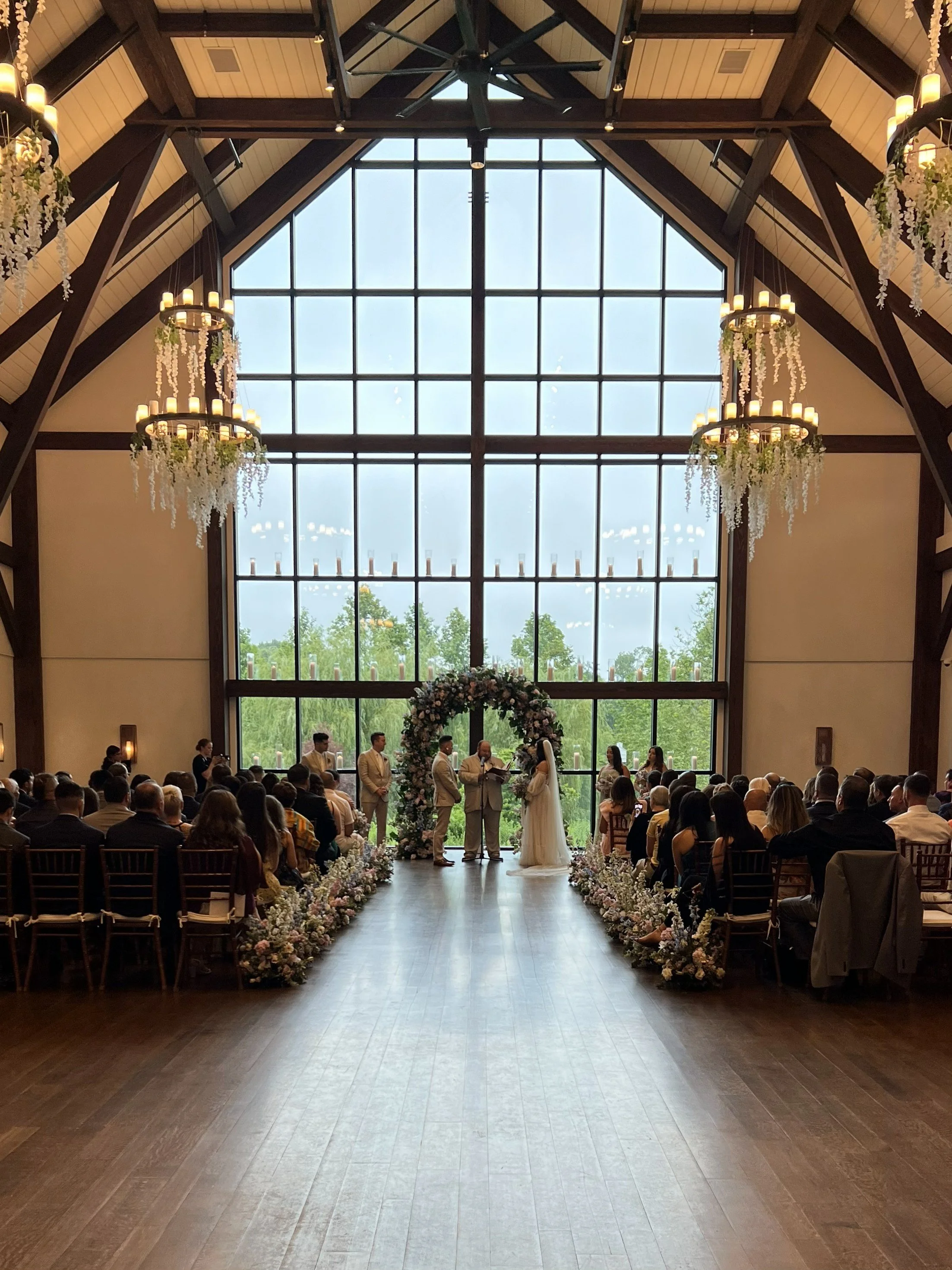 Indoor wedding ceremony with bride and groom standing under a floral arch, officiant reading, guests seated on both sides, large glass window in the background, and chandeliers hanging from a wooden ceiling.